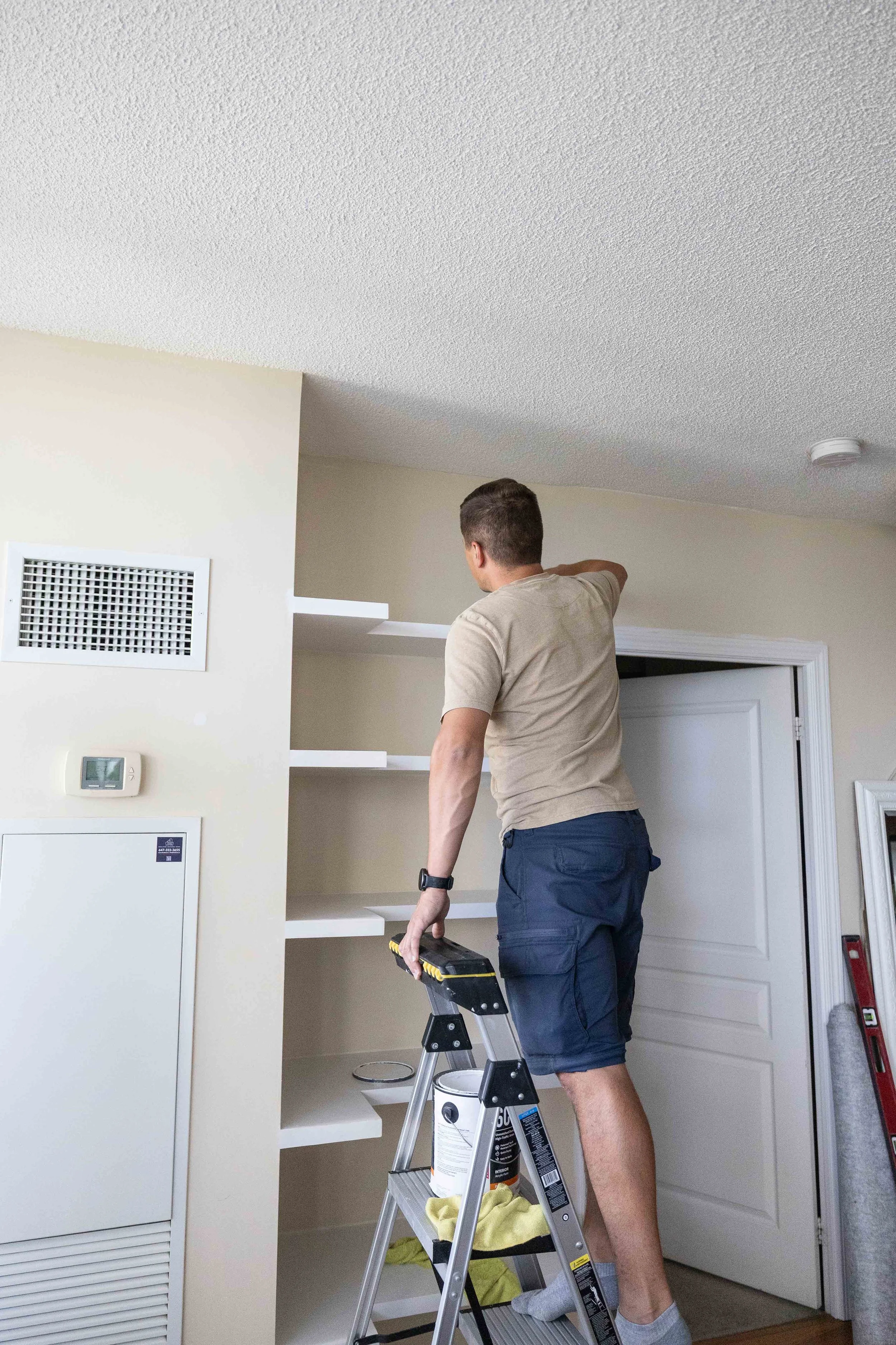 A man standing on a step stool, painting the inside of a white wooden cabinet in a room with beige walls and a white door.