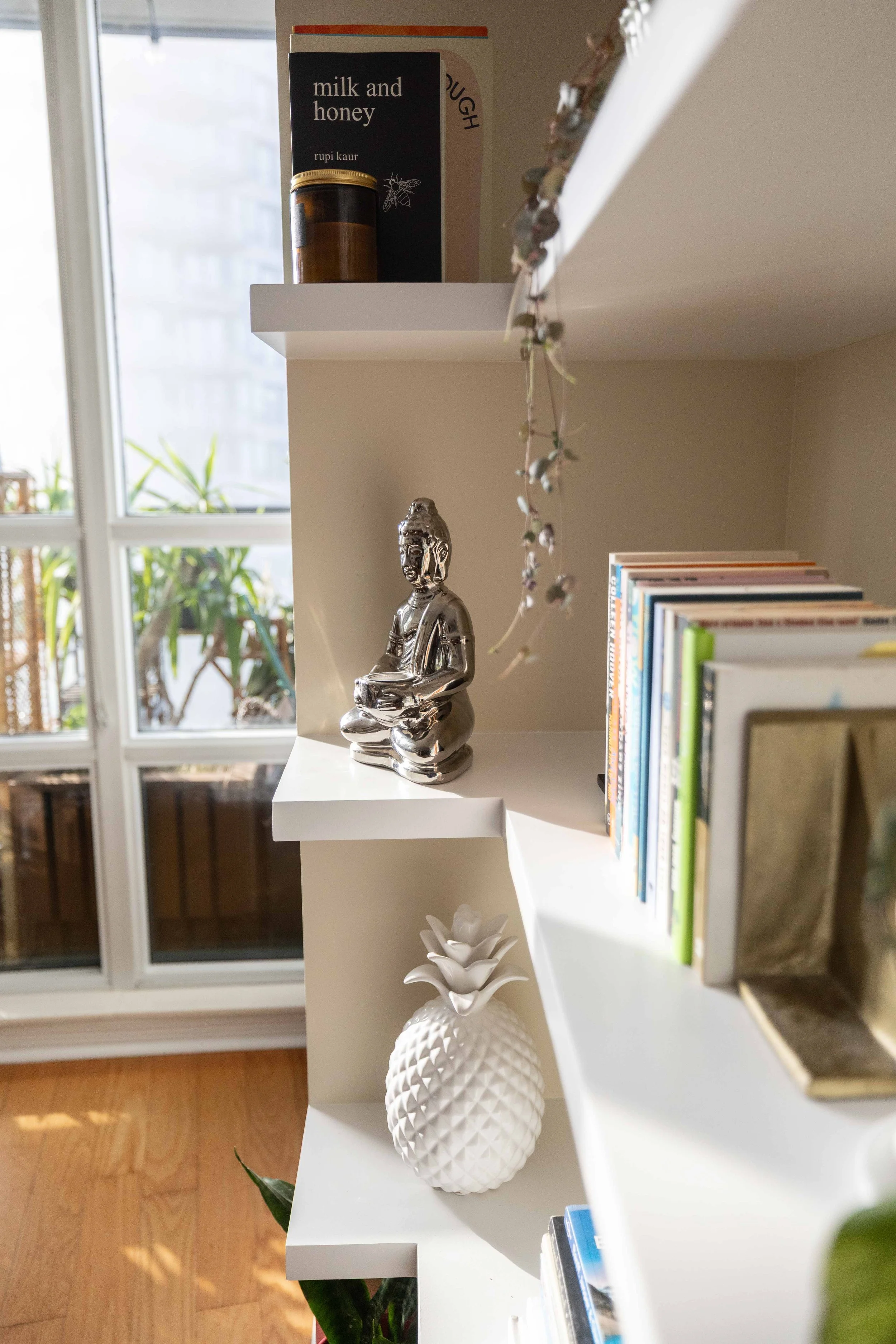 A white shelf with a black book titled 'milk and honey' by Rupi Kaur, a jar, a silver Buddha statue, a white pineapple-shaped ornament, and books on the right. A window behind the shelf lets in natural light, with plants outside.