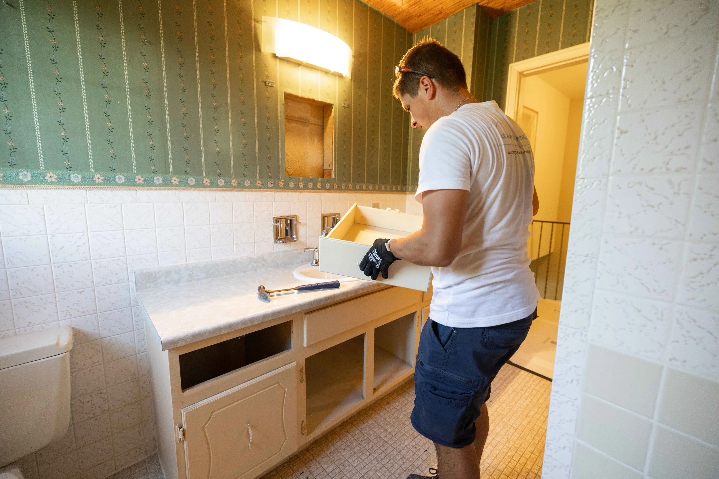 A man wearing a white t-shirt, shorts, and gloves is installing or working on a bathroom sink vanity. He is in a bathroom with vintage green wallpaper, white tiled wall, and a single light fixture above the mirror opening.