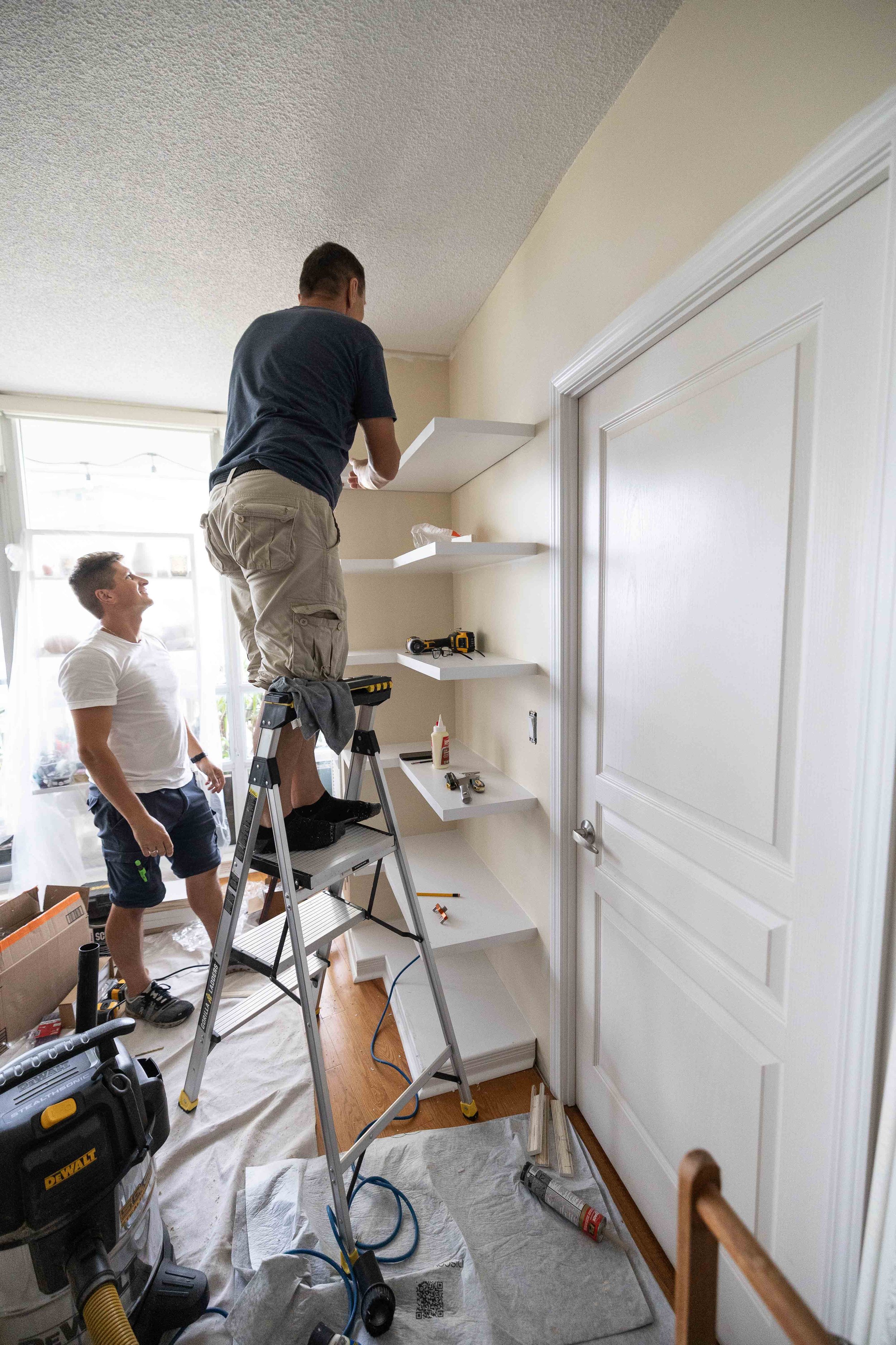 Two men installing white floating shelves on a beige wall tall ladder, power tools, and supplies on the floor.
