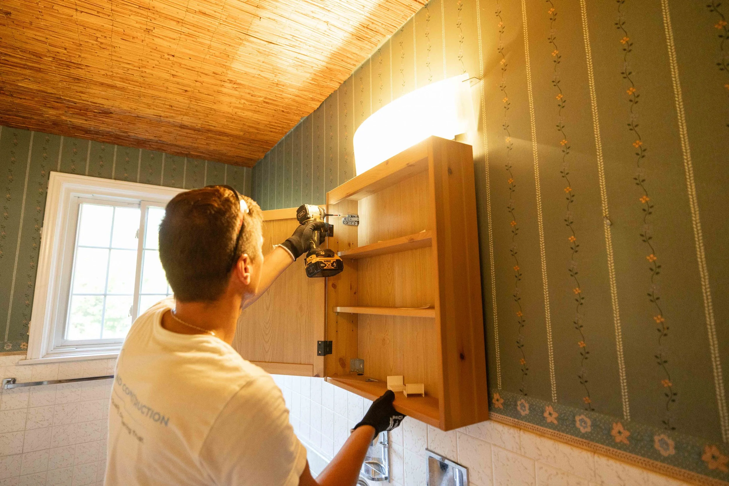 Person using a cordless drill to assemble a wooden piece of furniture next to a window in a room with floral patterned wallpaper and wooden ceiling.