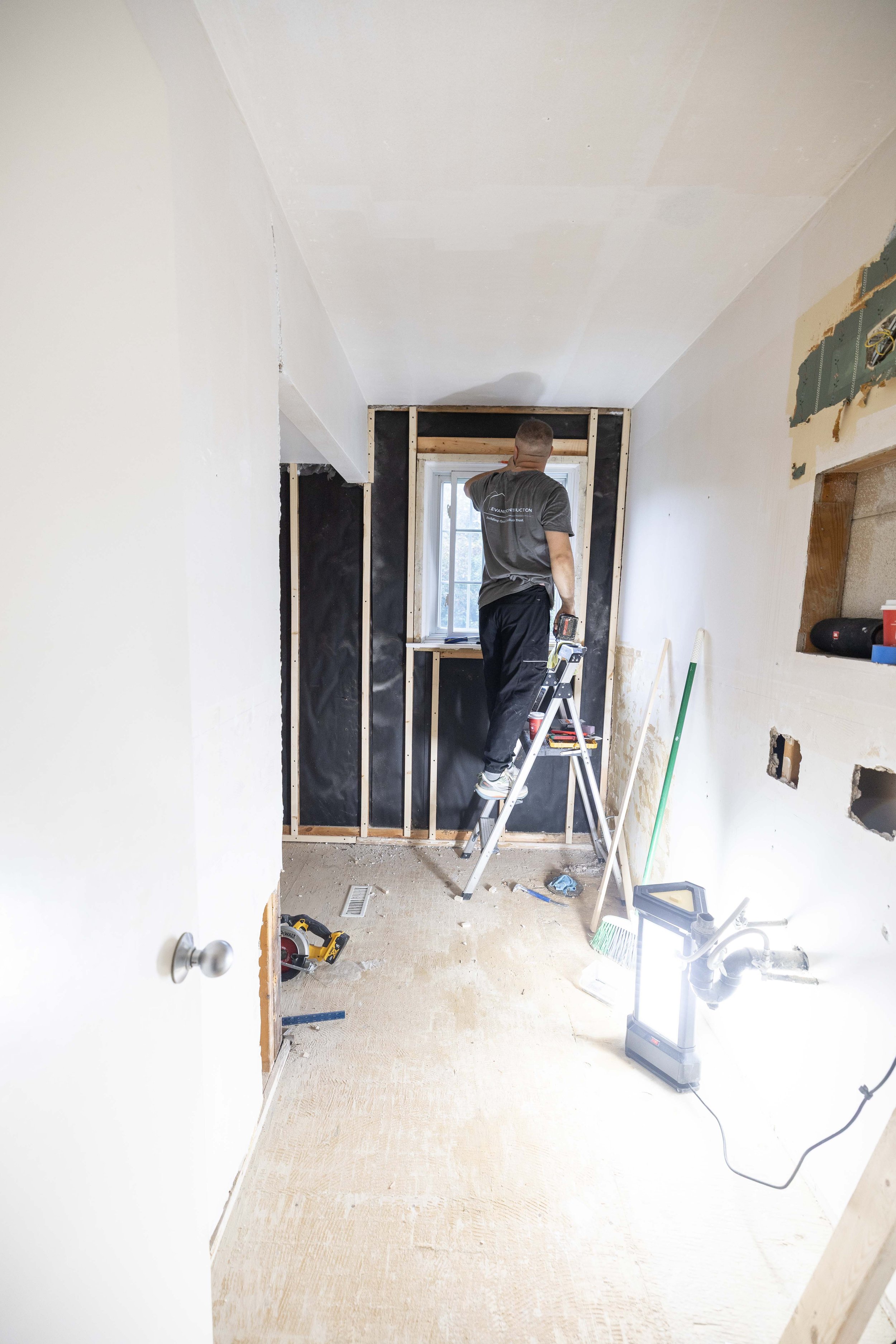 A man is working on a bathroom renovation, installing framing around a window, with tools and construction materials scattered on the floor.