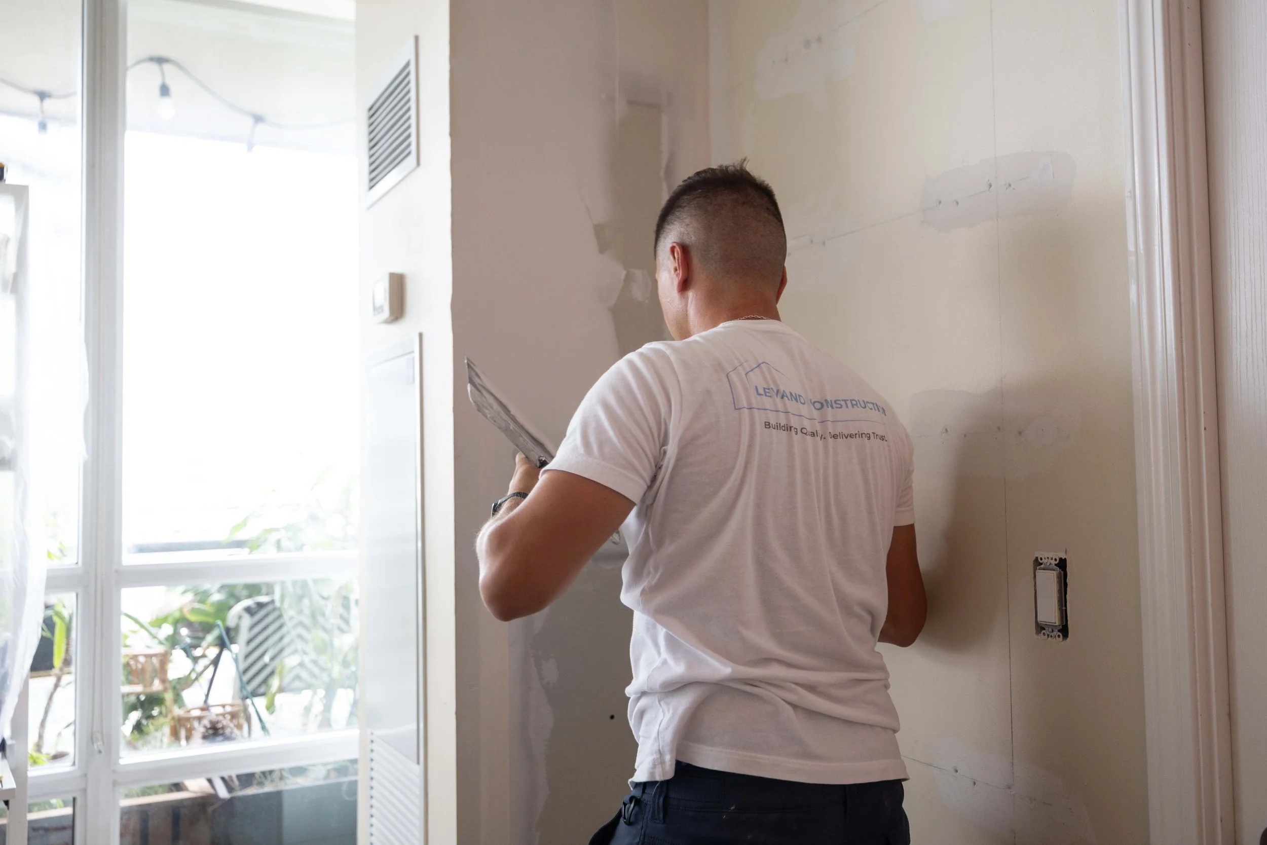 A man with a short haircut wearing a white T-shirt with blue print, standing inside a room, holding a tool, with an unfinished wall and a large window in the background.