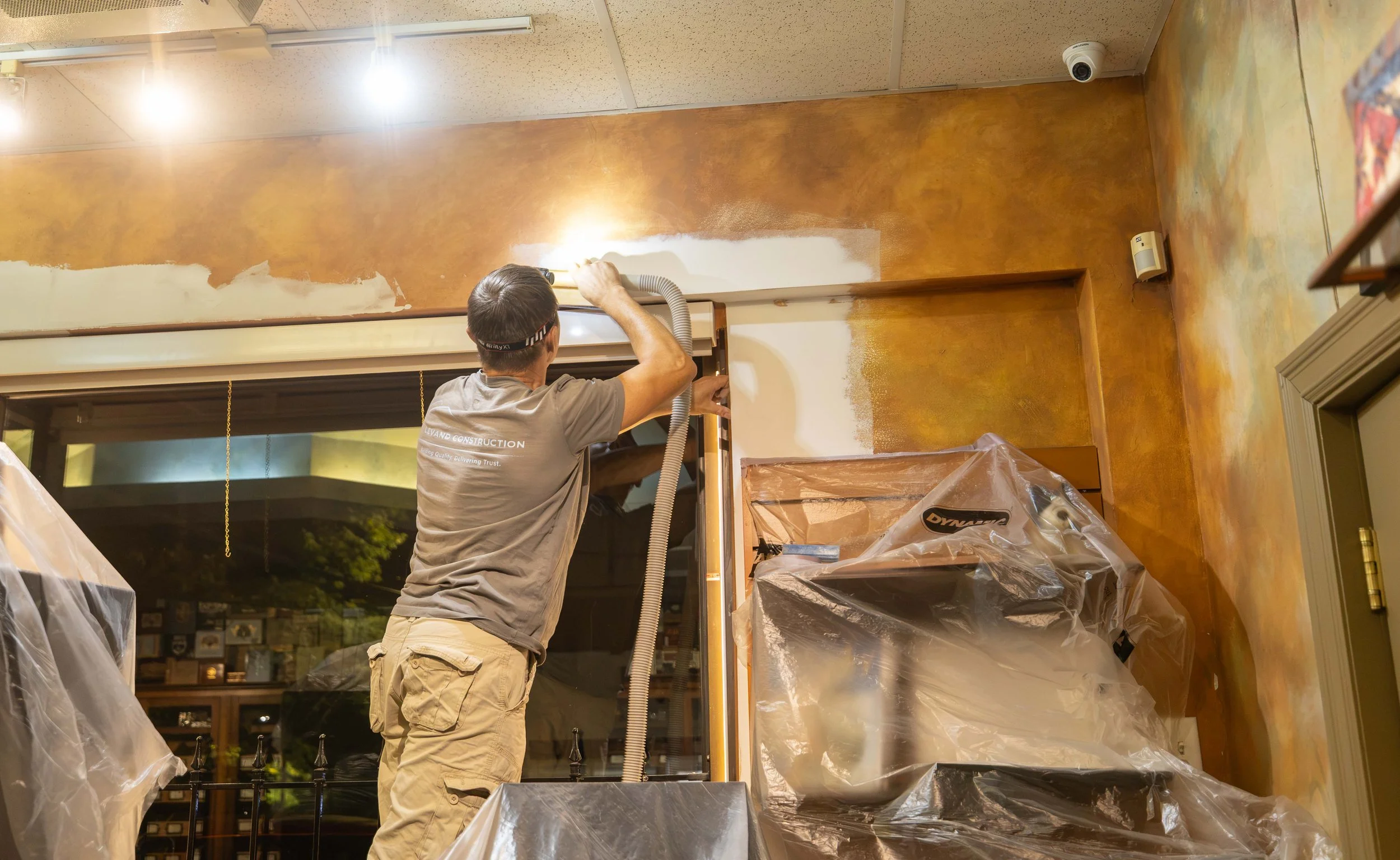 A man is painting a wall inside a room, using a spray gun connected to a hose. The room has a brown textured wall, and the man is wearing a gray t-shirt and tan shorts. Plastic covers and equipment are visible around the area.