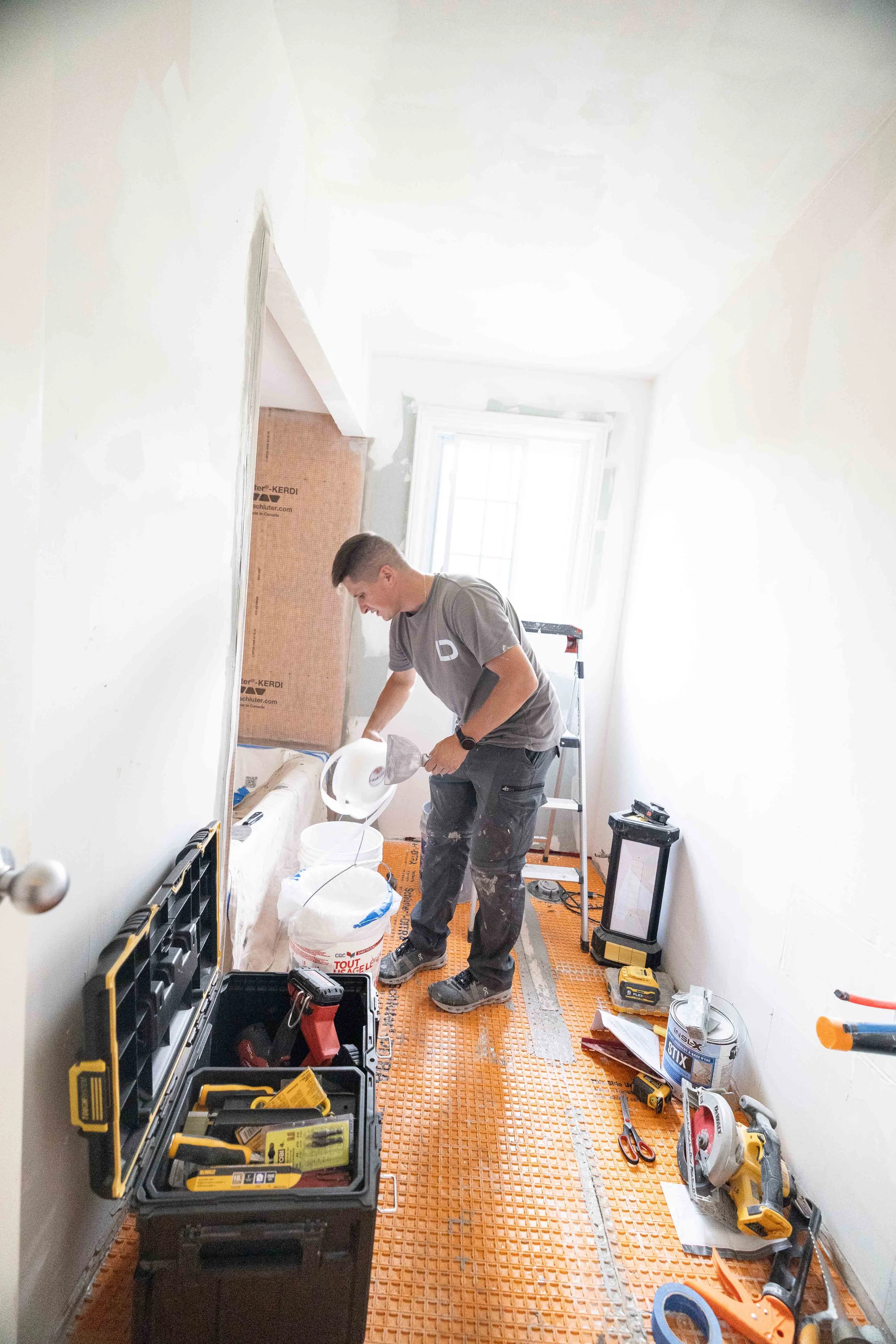 A man working on a home renovation inside a room, surrounded by tools, construction materials, and equipment. Sunlight streams through a window.