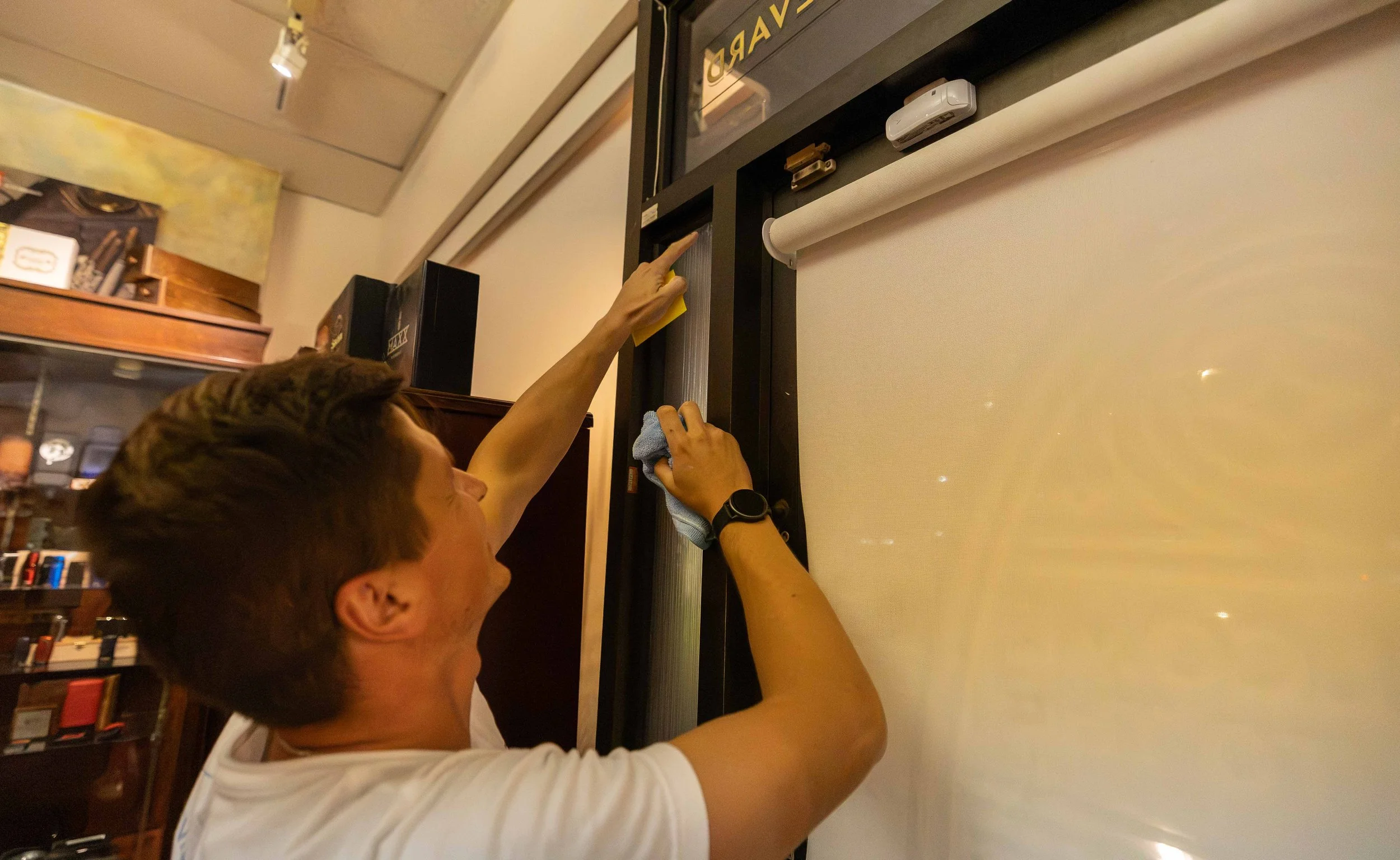 Two men are cleaning a film projector screen in a room with bookshelves. One man is wiping the screen while the other is holding a yellow cloth. The room has a cozy, organized atmosphere.