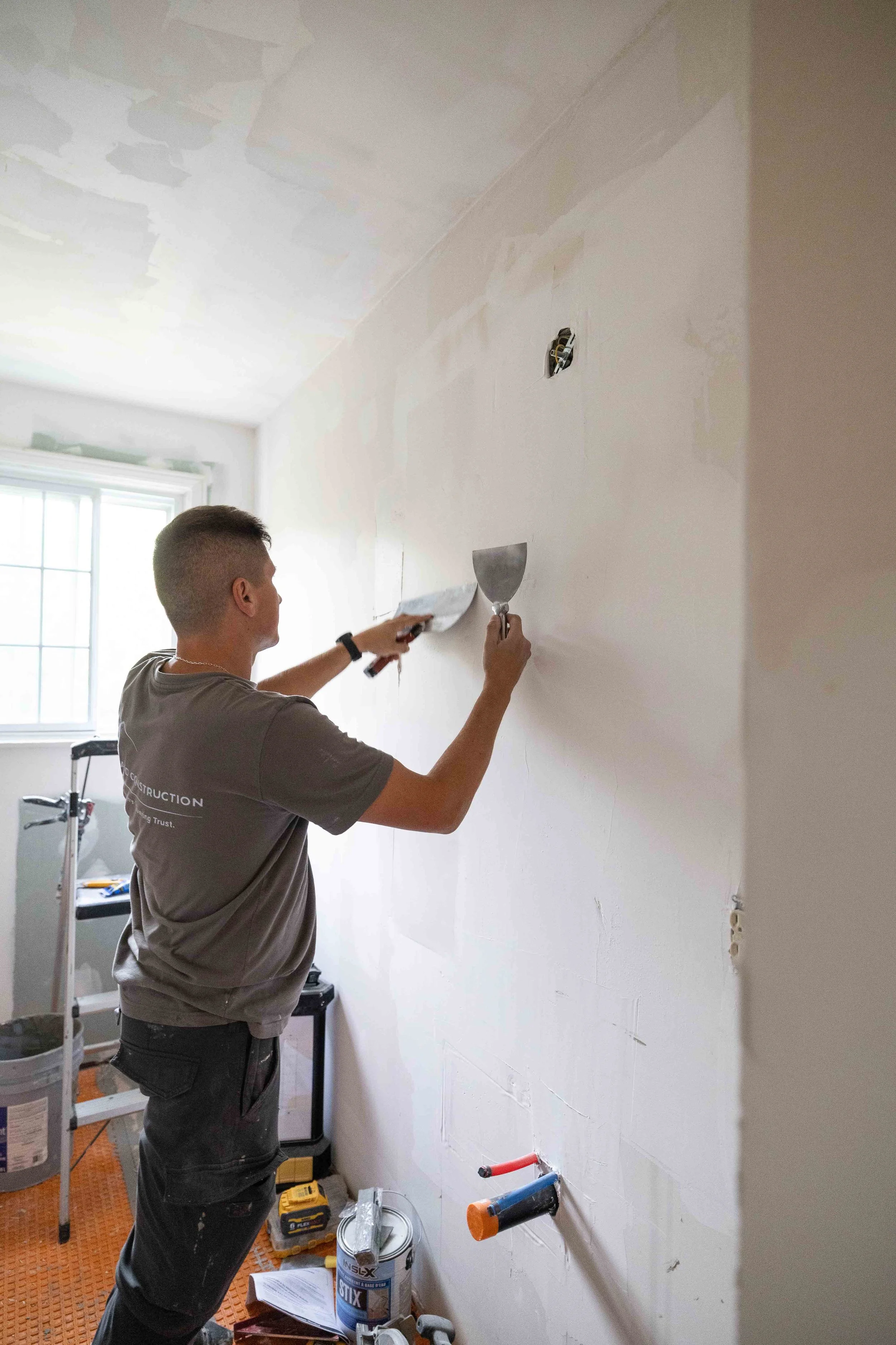 A man is installing or repairing electrical wiring in a room, working on a wall socket with exposed wires, surrounded by construction tools and supplies.