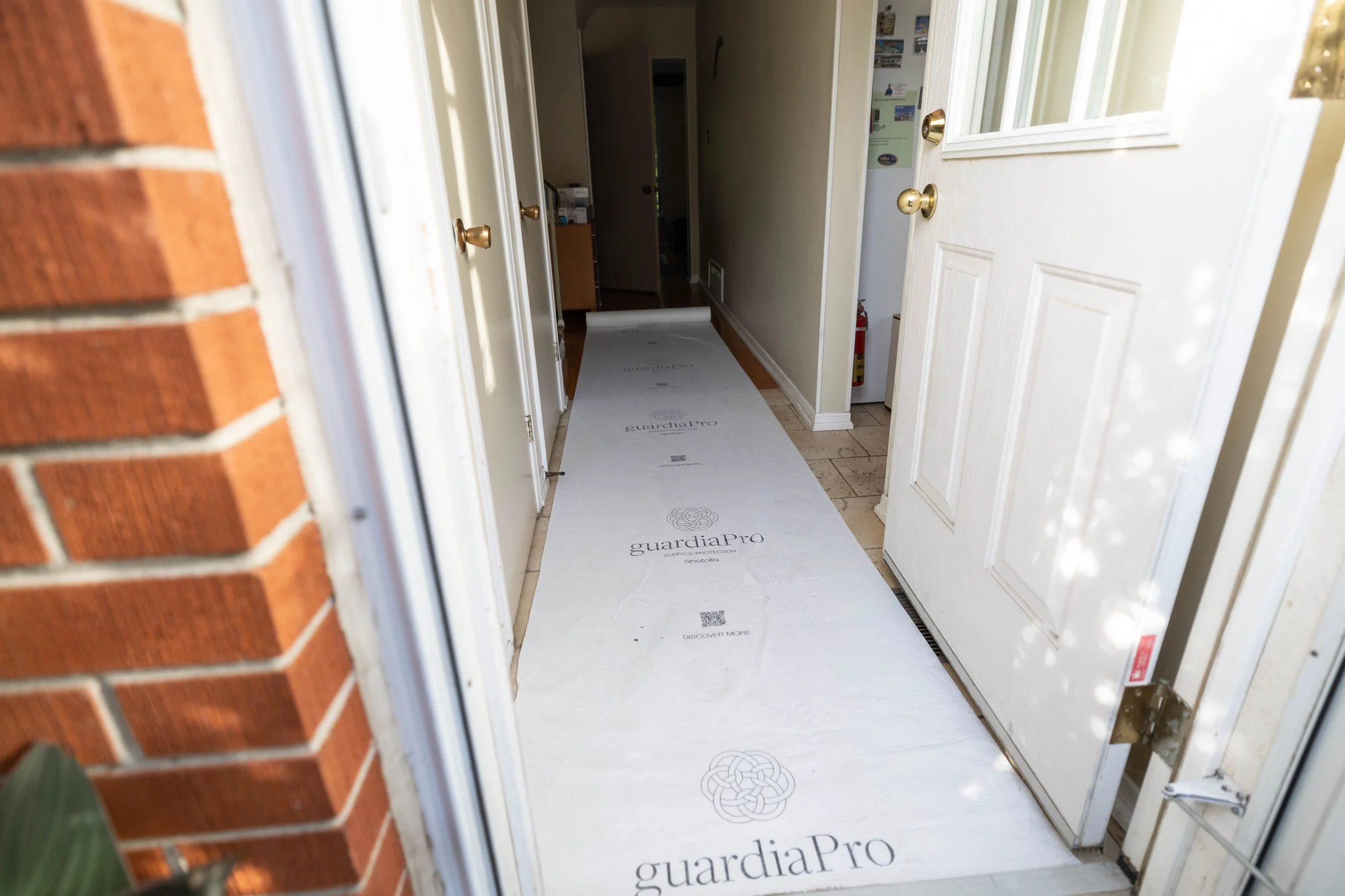 View of a house entrance showing a door open to reveal a hallway with cardboard floor protection labeled 'guardiaPro'. The hallway has light-colored walls and a fire extinguisher at the end.