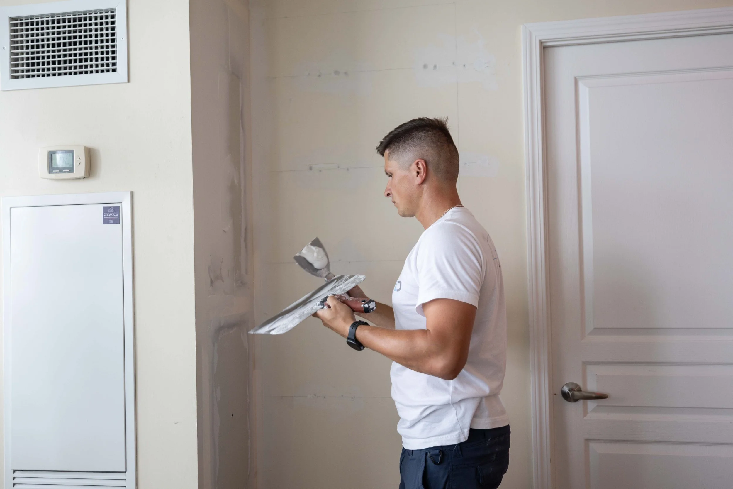 A man wearing a white t-shirt and dark pants is painting a wall with a paintbrush and tray. The wall has patches of primer or paint and is near a white door and wall-mounted thermostat.
