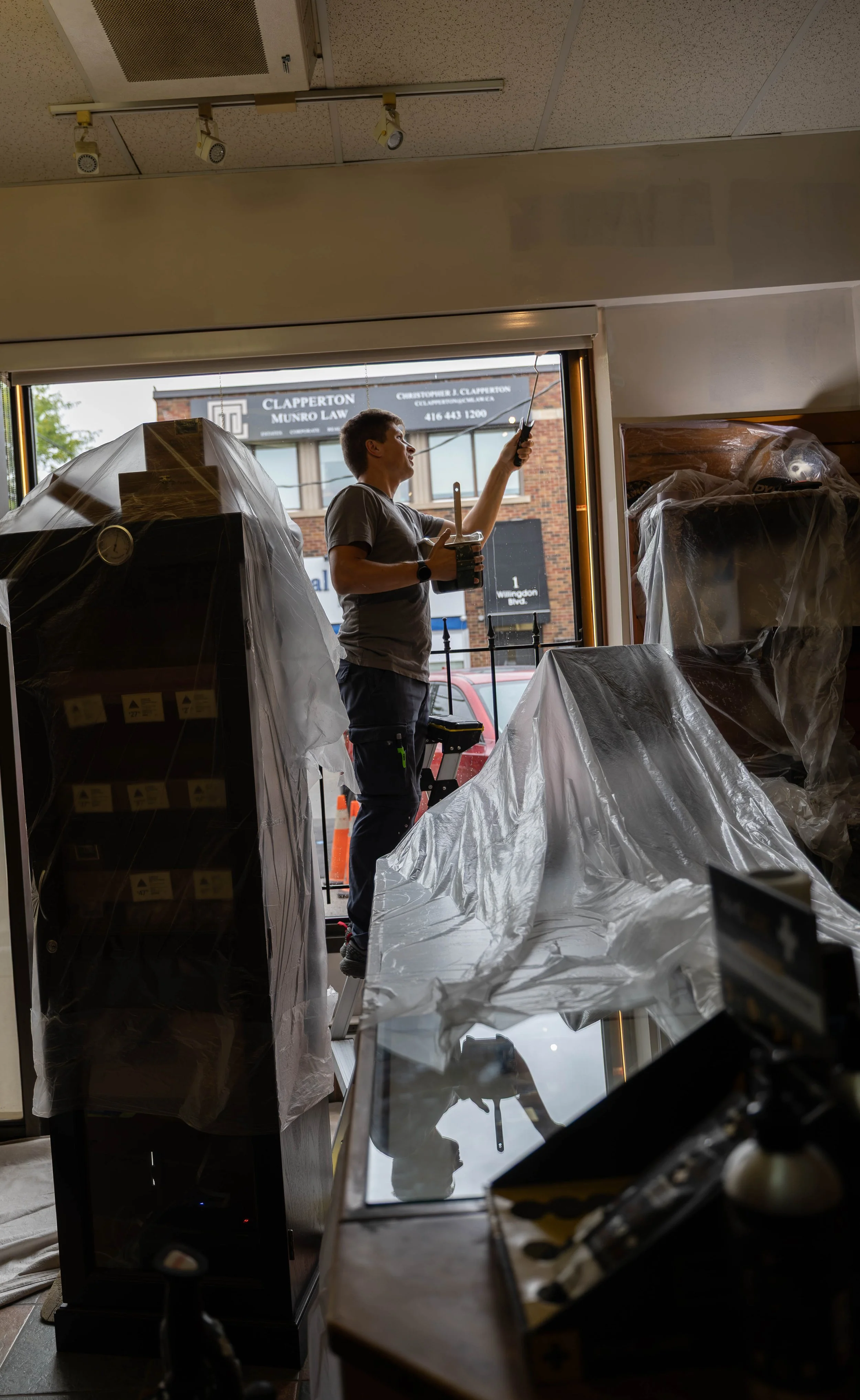 A man standing on a small step ladder, inside a store, using a screwdriver to work on an object near the ceiling, with plastic-covered shelves and a window showing a building and street outside.