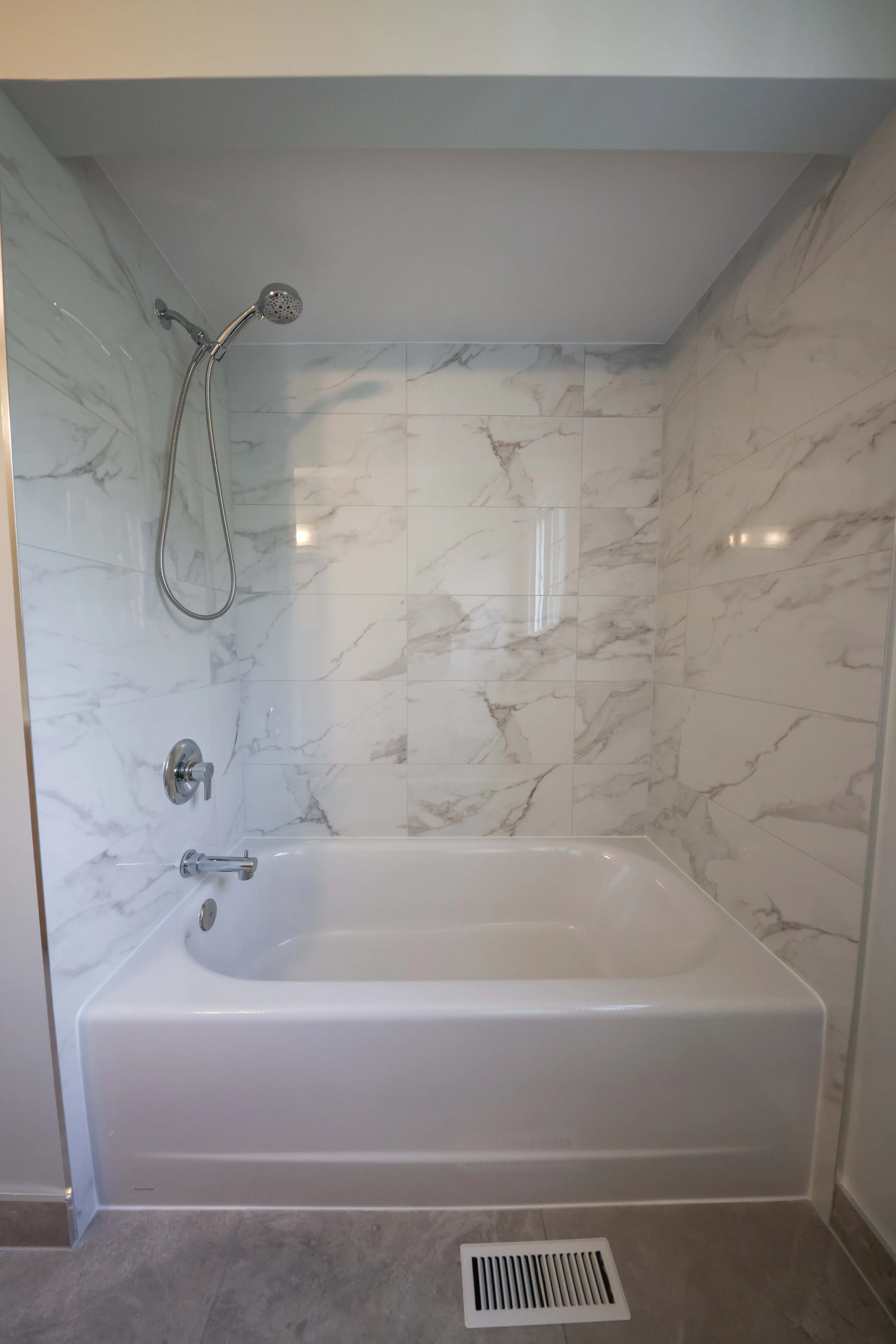A bathroom with a white bathtub, marble tile walls, a handheld showerhead, and a floor vent.