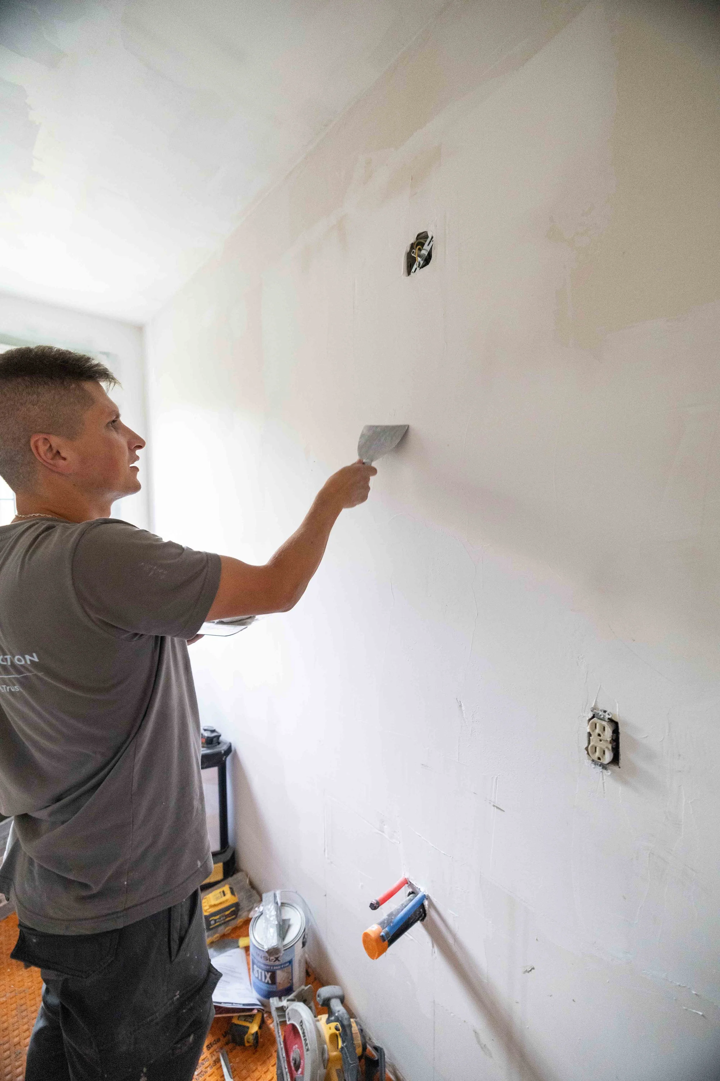 A man in gray t-shirt and black pants working on a wall, using a putty knife. The wall has electrical outlets and exposed wiring. Tools and materials like a bucket, power tools, and a level are on the floor.