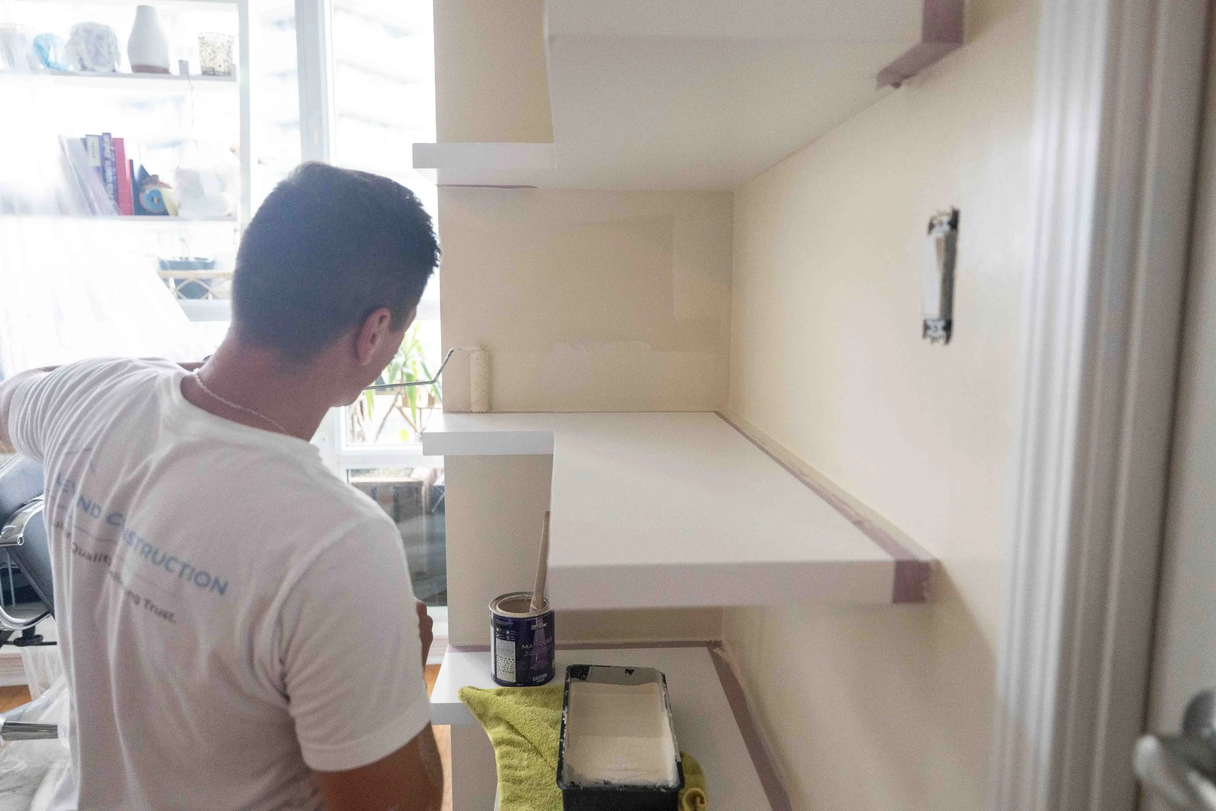 Person painting a white shelf in a room with a partially painted wall.