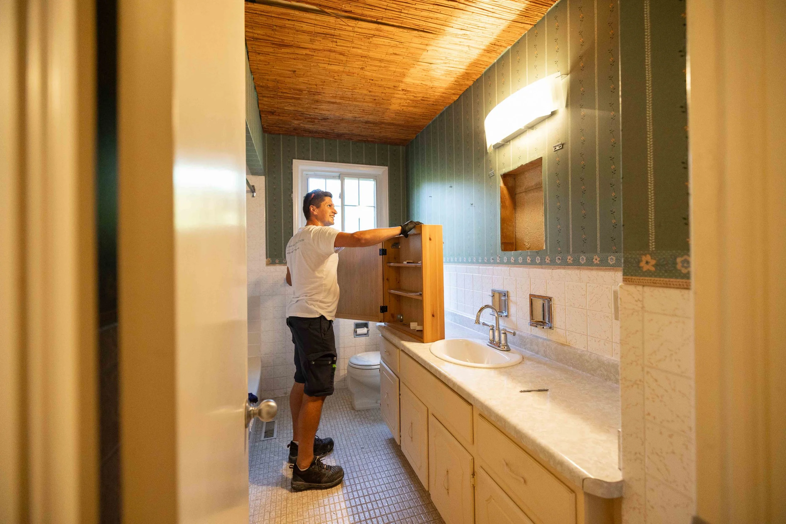 A man in a white T-shirt and black shorts working on a bathroom cabinet above the sink, with green wall wallpaper, wood ceiling, and a window in the background.