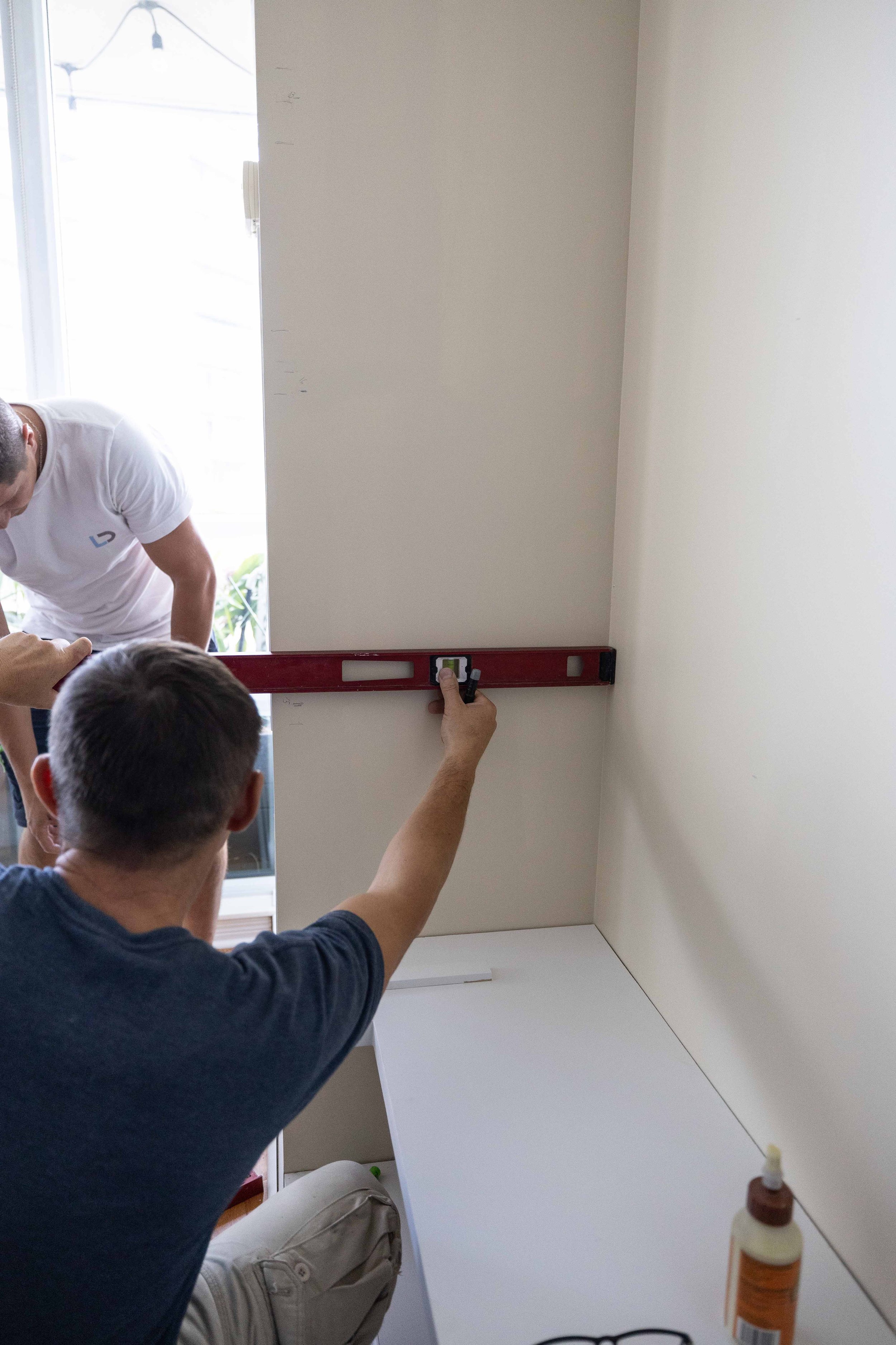 A group of people is installing or working on a large mirror or photo frame on a wall in a room with white walls. One person is using a level to ensure proper alignment, while others are observing and assisting. There is natural light coming from a d