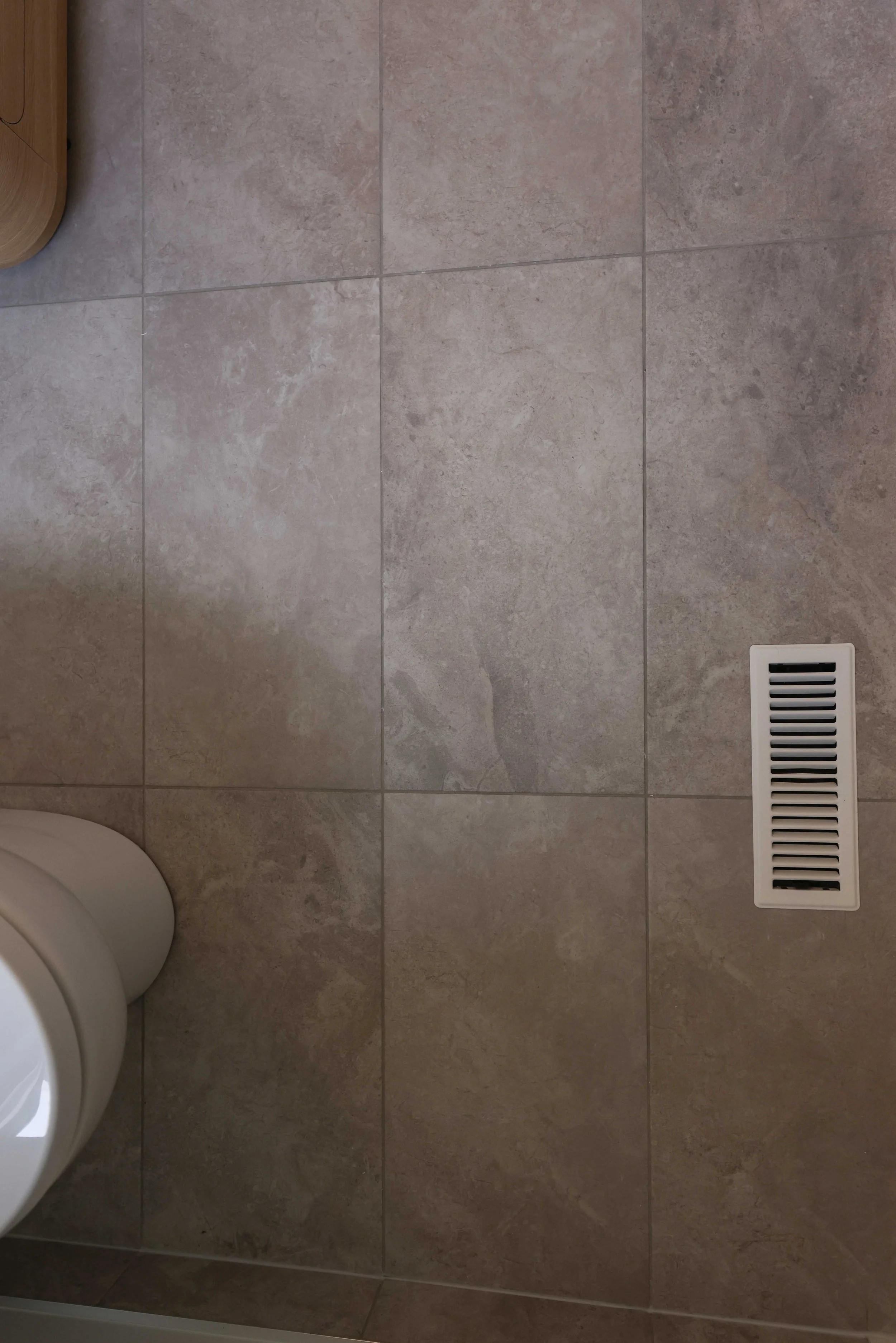 Bathroom floor with beige tiles, a white ventilation grill, and part of a white toilet visible in the bottom left corner.