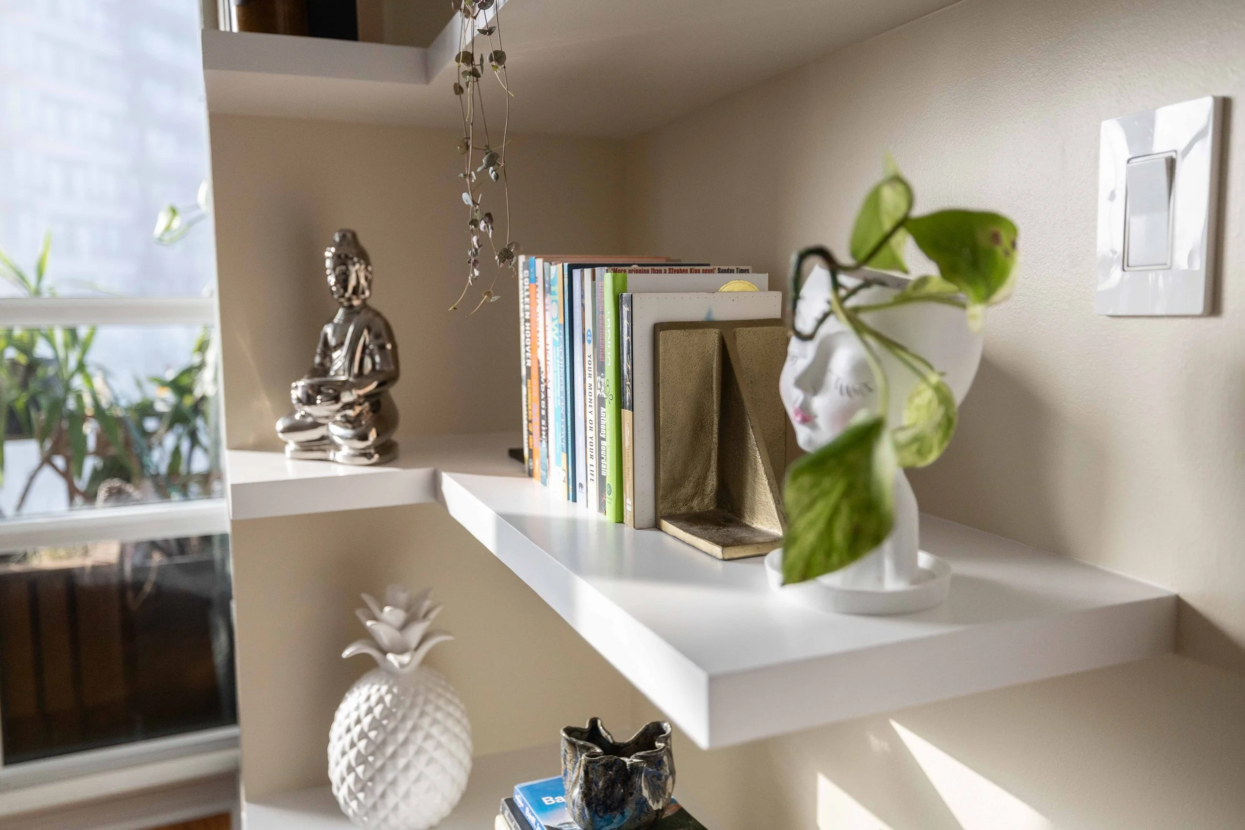 White bookshelf with decorative objects, a collection of books, and a hanging plant, with a window in the background.