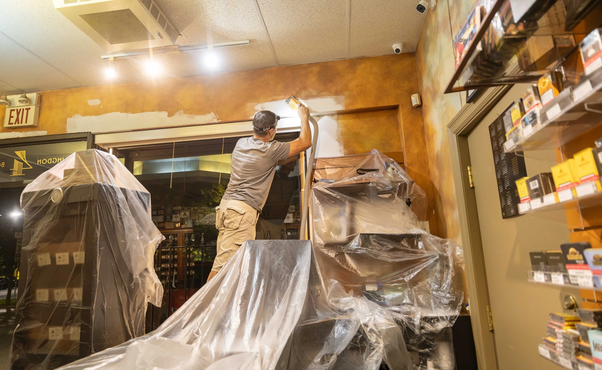 A man is painting a wall inside a store with a paint roller. The area is partially covered with plastic sheets, and the ceiling has bright lights. There is an exit sign on the wall and shelves with products.