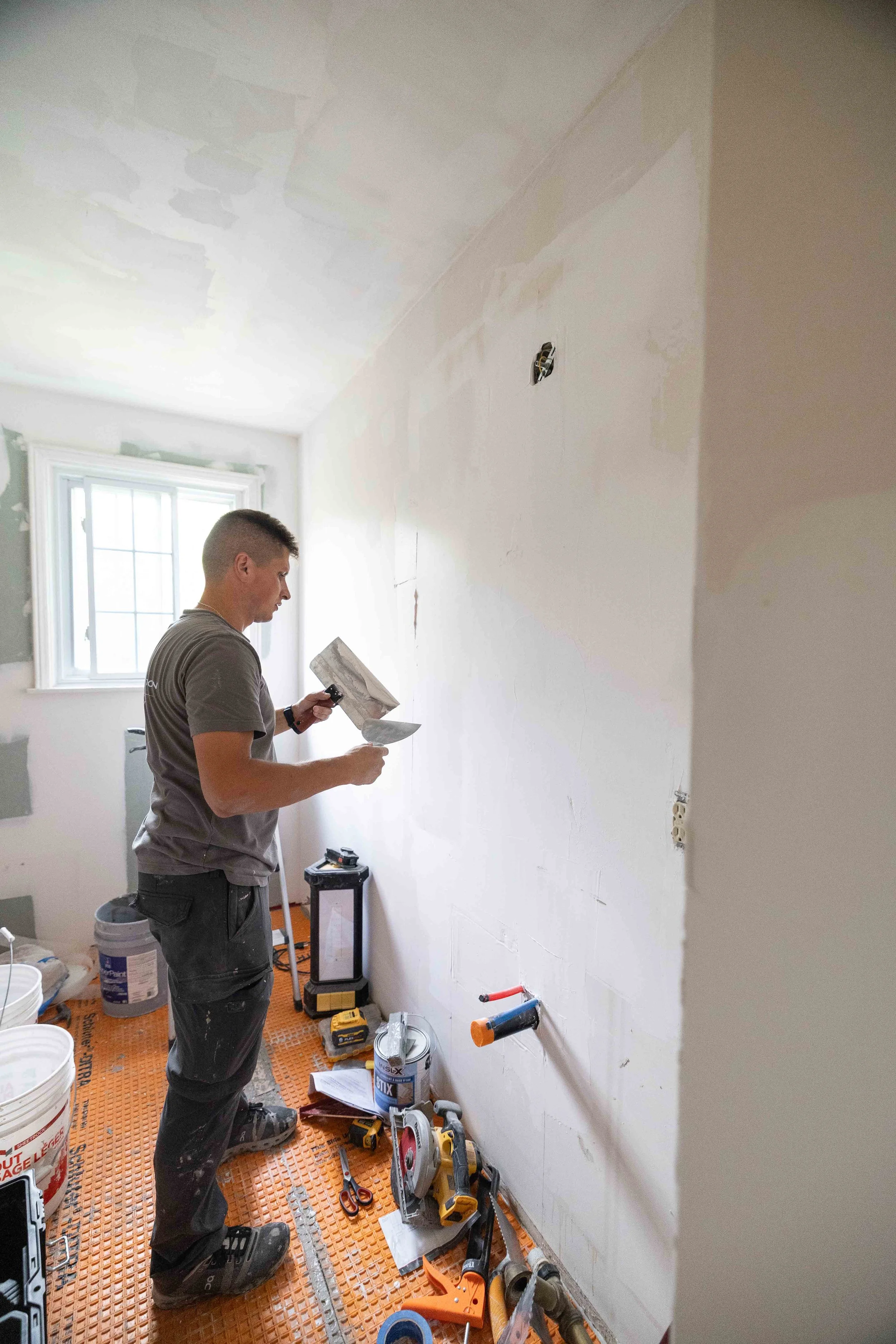 A man wearing a gray T-shirt and black pants working on drywall in a room under renovation, with various tools, paint, and construction materials scattered on the orange floor covering.