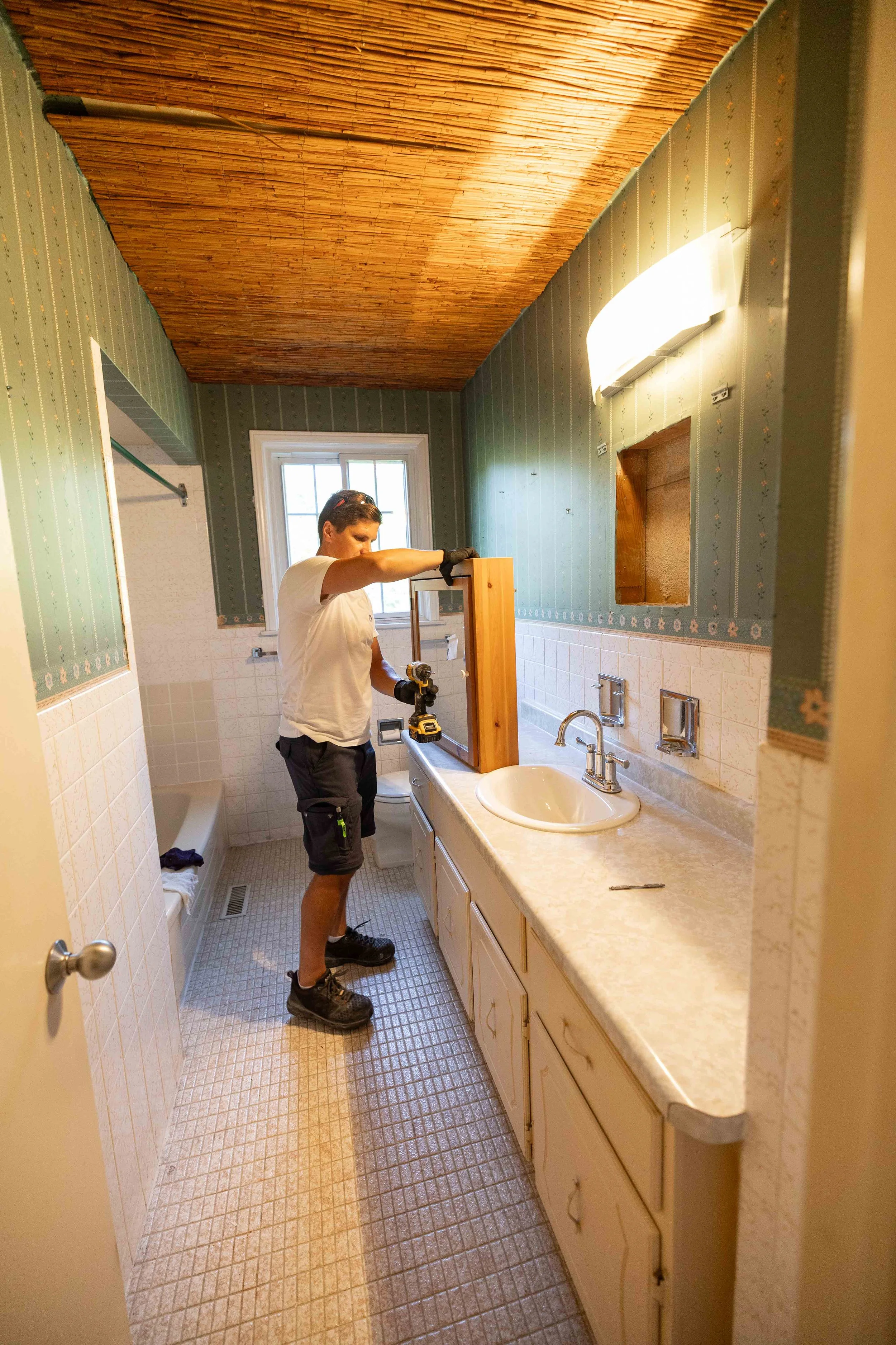 A man installing or repairing a wooden cabinet in a bathroom with a granite countertop. The bathroom features green wallpaper with small floral patterns, a window, and a tiled floor. The man is wearing a white t-shirt, black shorts, and work boots, a