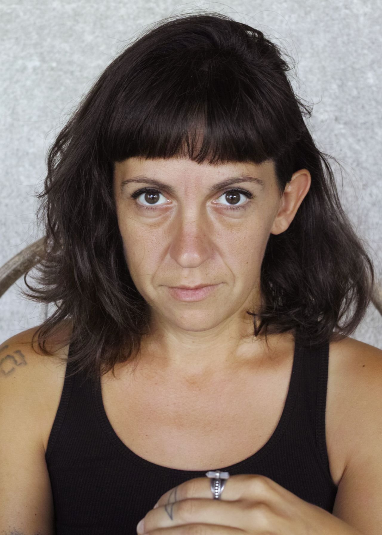 Portrait d'une femme aux cheveux courts, foncés, et aux yeux marron, portant un débardeur noir, assise contre un mur gris, avec tatouages visibles sur l'épaule et le bras gauche, portant une bague à la main droite.