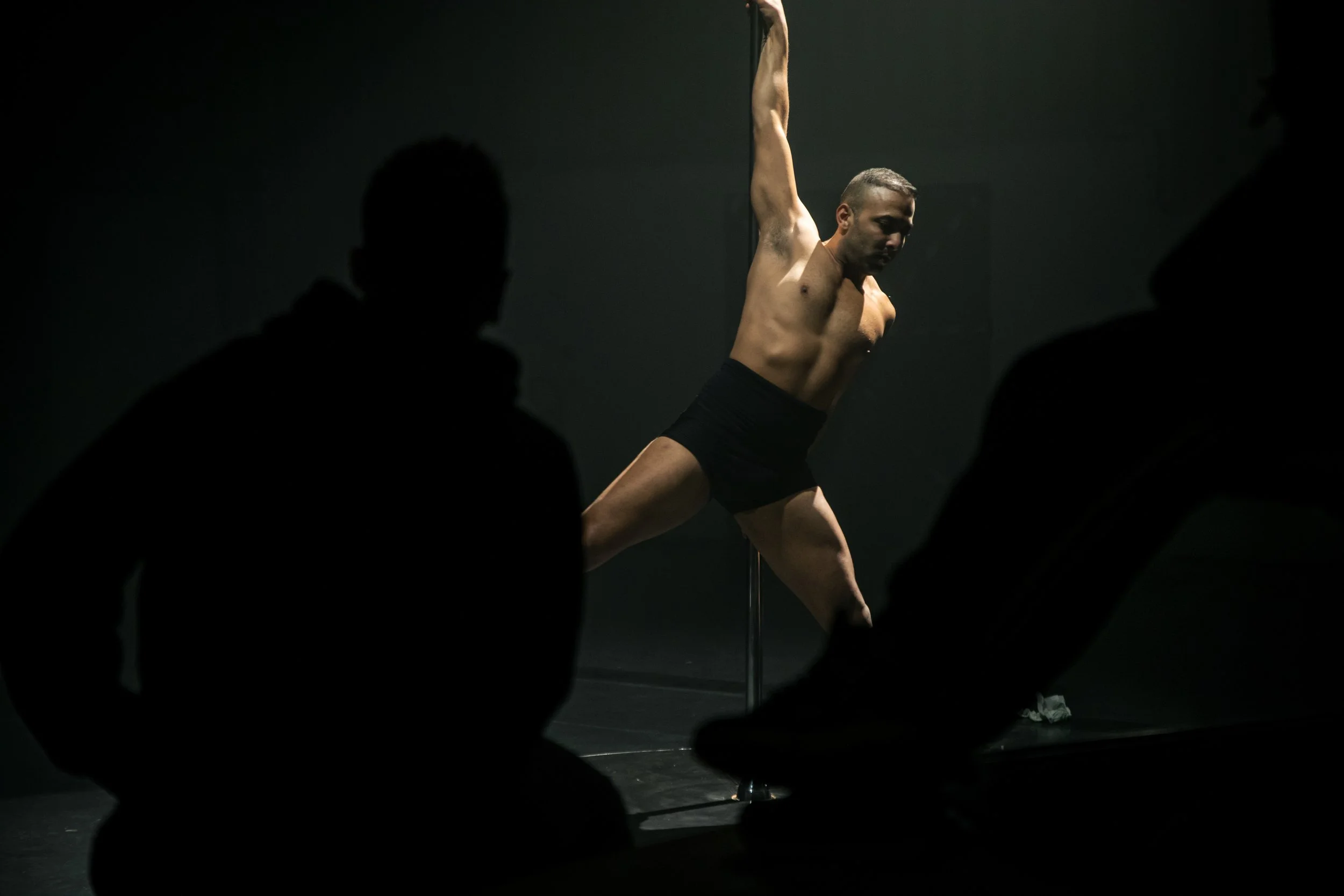 Un homme en maillot noir effectue une performance de pole dance dans une pièce sombre, avec des silhouettes d'ovateurs en arrière-plan.