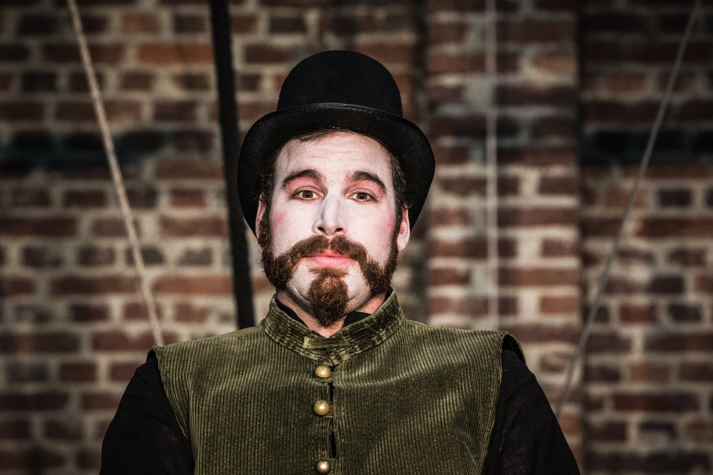 Un homme avec une barbe, des joues rouges et portant un haut-de-forme noir, un costume vert à boutons dorés, se tenant devant un mur en briques.