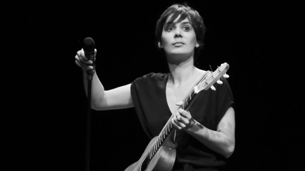 Une femme chante et joue de la guitare sur scène, tenant un micro dans la main droite. En noir et blanc, elle a les cheveux courts et porte un vêtement sombre.
