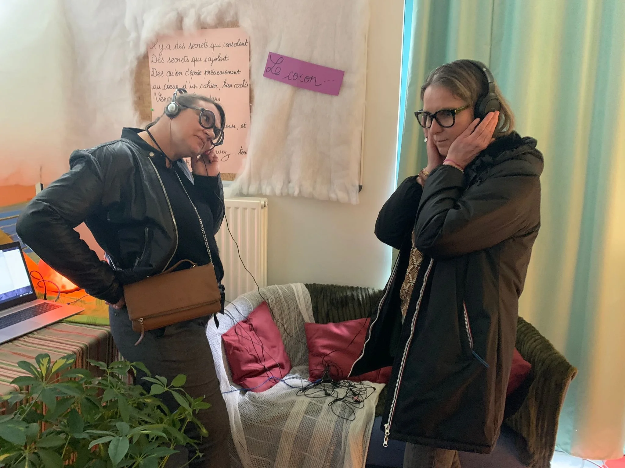 Deux personnes, un homme et une femme, portent des casques audio, semblent écouter de la musique ou participer à une activité audio. La pièce semble être un salon avec un canapé, des coussins, et un tableau avec du texte écrit en arrière-plan. La femme porte des lunettes et une veste noire, l'homme porte une veste en cuir.