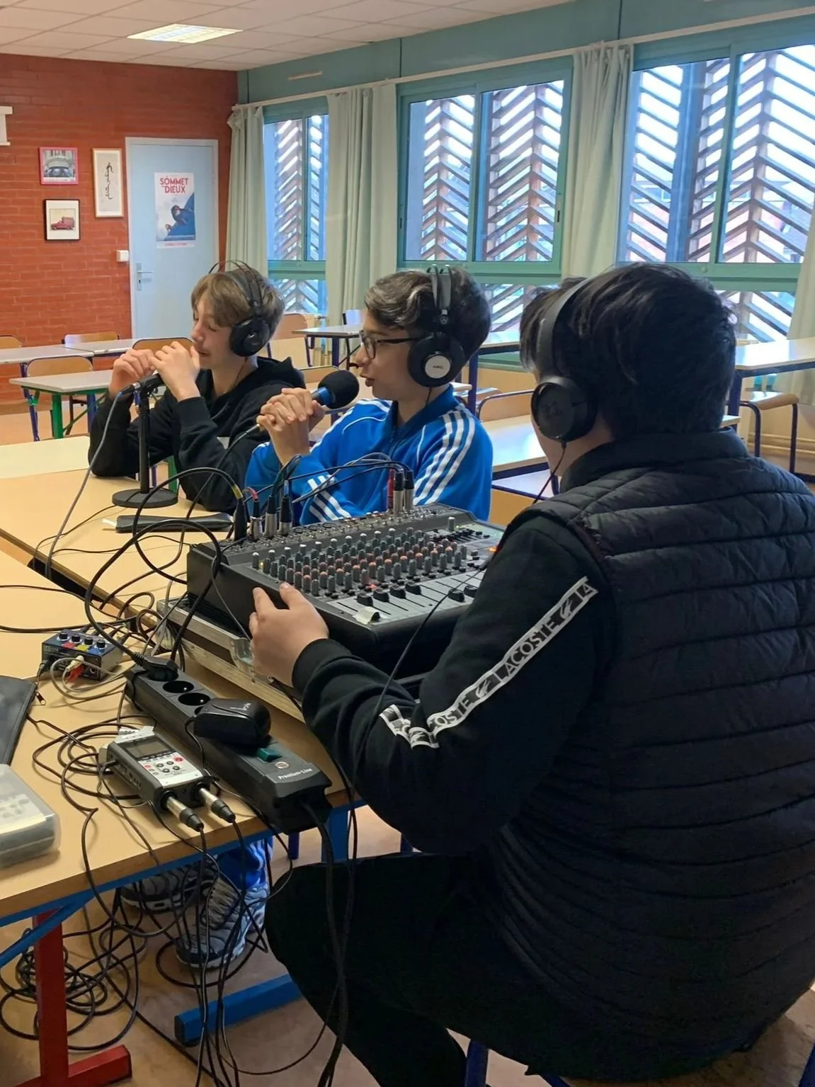 Trois jeunes hommes participent à une séance de radio ou d'enregistrement audio dans une salle, avec du matériel sonore, et portent des casques audio.