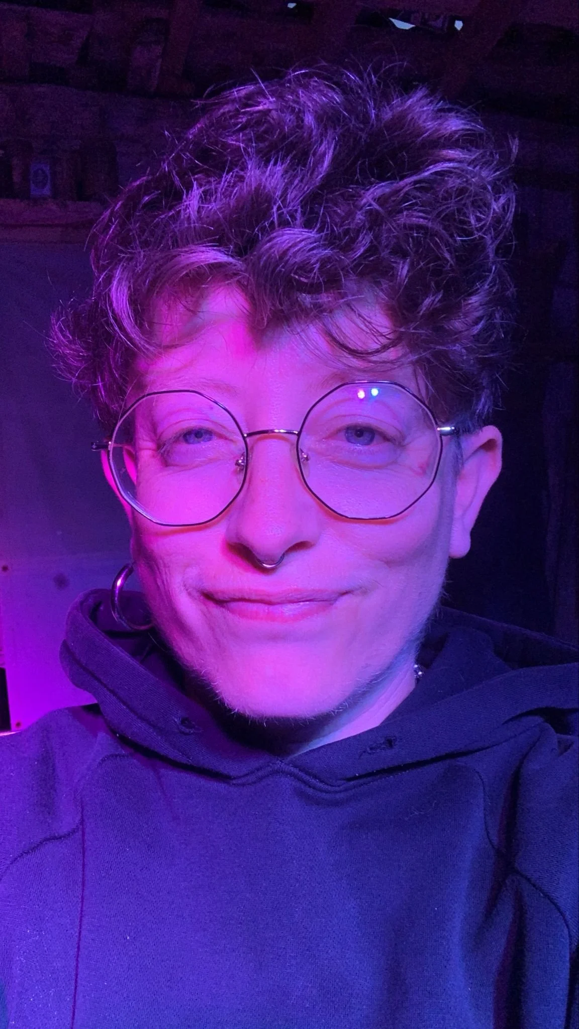 Portrait d'une personne avec des lunettes rondes, boucle d'oreille en cercle, sourire léger, coloration violette et lumière bleue, cheveux frisés, portant un sweat noir.
