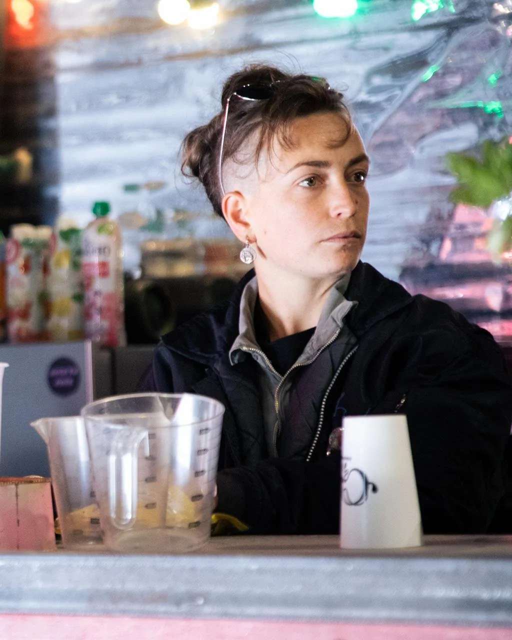 Une femme assise derrière un comptoir, regardant vers la droite, avec des bouteilles et des éléments décoratifs en arrière-plan.