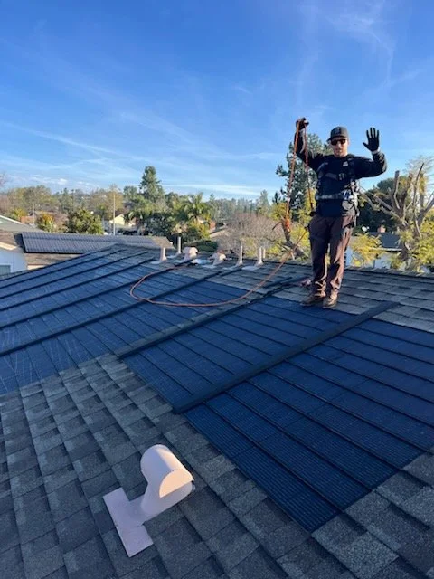 A man standing on a residential roof with solar panels, holding a safety rope, wearing a harness, and waving at the camera.