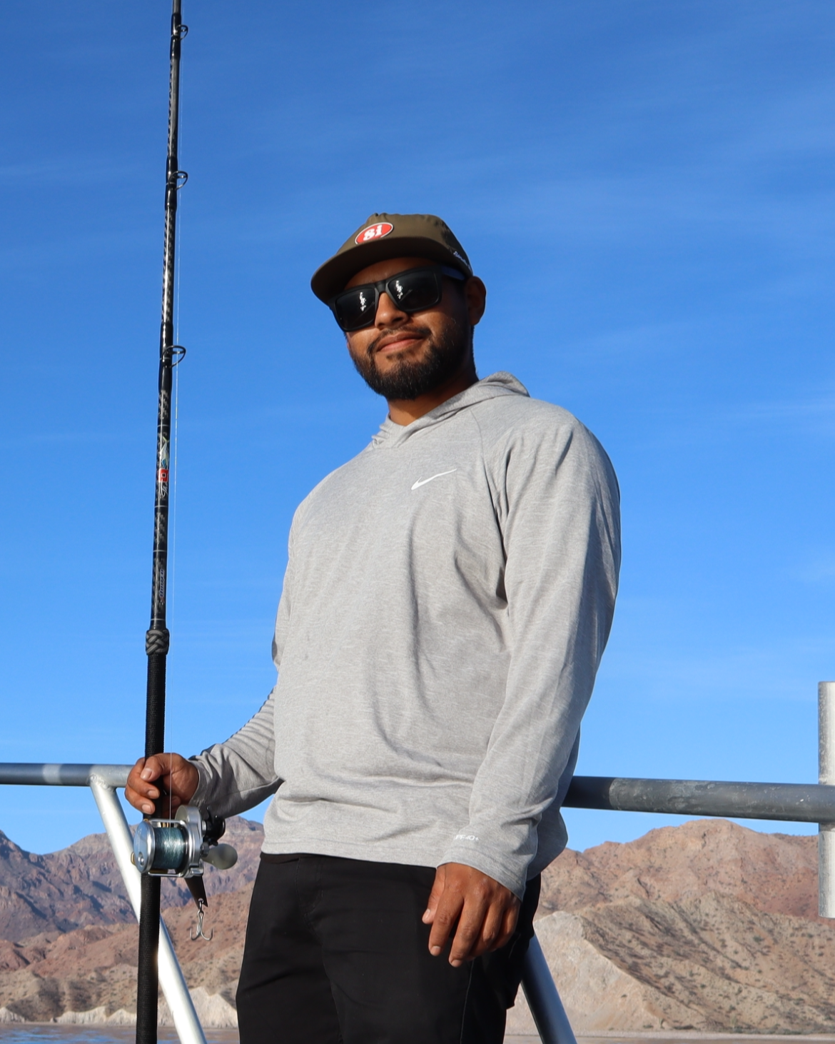 Man standing outdoors holding a fishing rod, wearing sunglasses, a gray Nike hoodie, and a baseball cap, with mountains and a clear blue sky in the background.