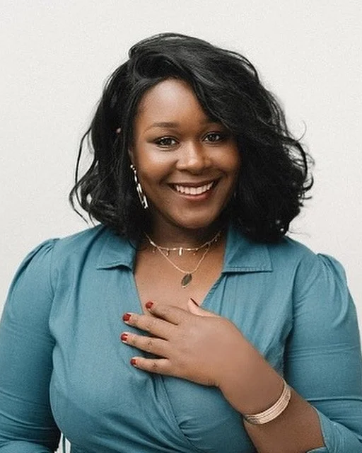 A woman with shoulder-length curly black hair, wearing a teal blouse, gold jewelry, and smiling with her hand on her chest, standing against a plain white background.