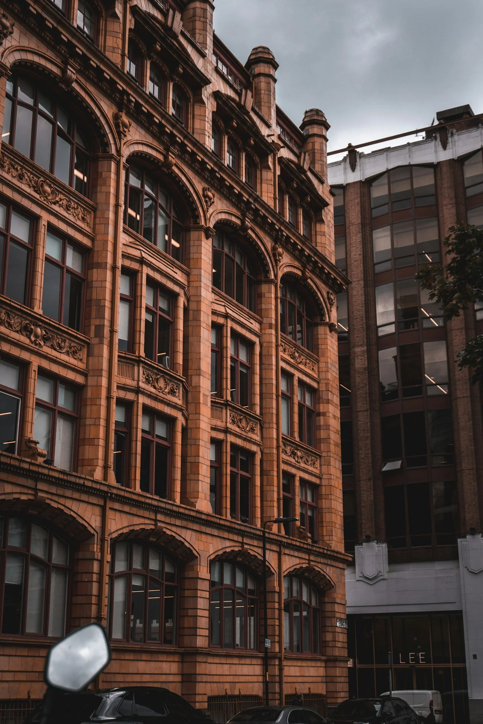 A large historic building with decorative brickwork and tall arched windows fronting a city street. Next to it is a modern glass and brick building. Vehicles are parked on the street, and a part of a car mirror is visible in the foreground. The sky is overcast.