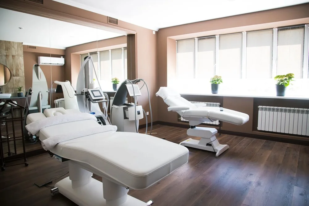 A medical or spa treatment room with a treatment bed, medical devices, and windows with potted plants.