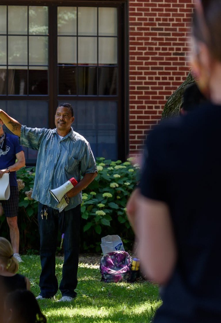A man is speaking to a group outdoors in front of a brick building with large windows, holding a megaphone and gesturing with his right hand, while several children and adults are listening.