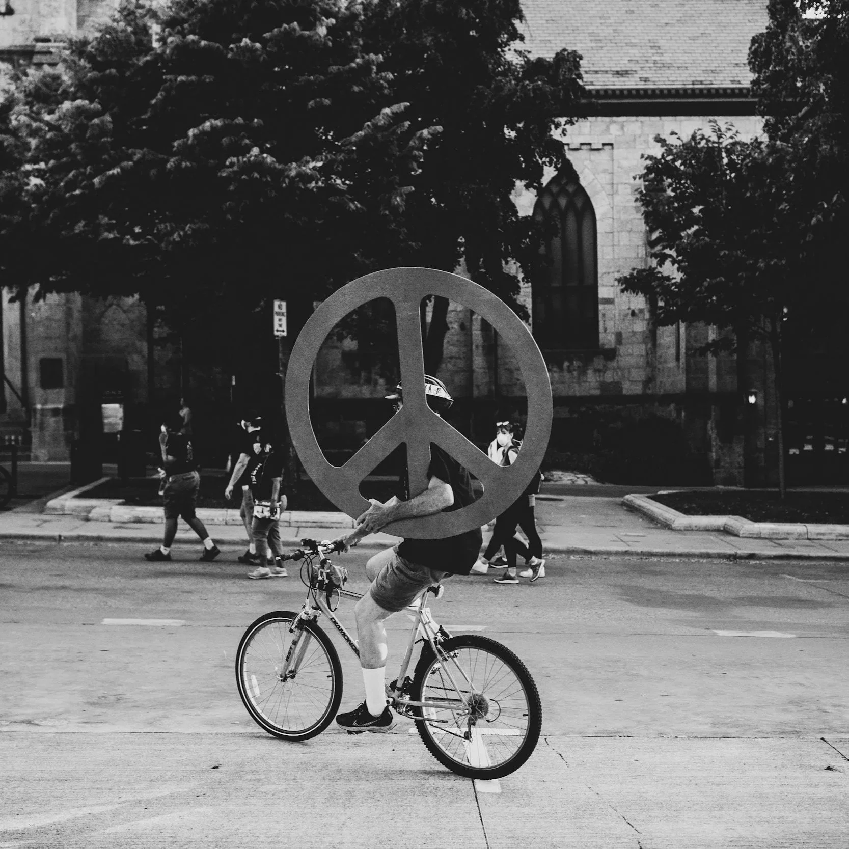 A person riding a bicycle on the street with a large peace sign prop in front of them, and a church building with pointed windows in the background. The person is holding the peace sign while riding the bike.
