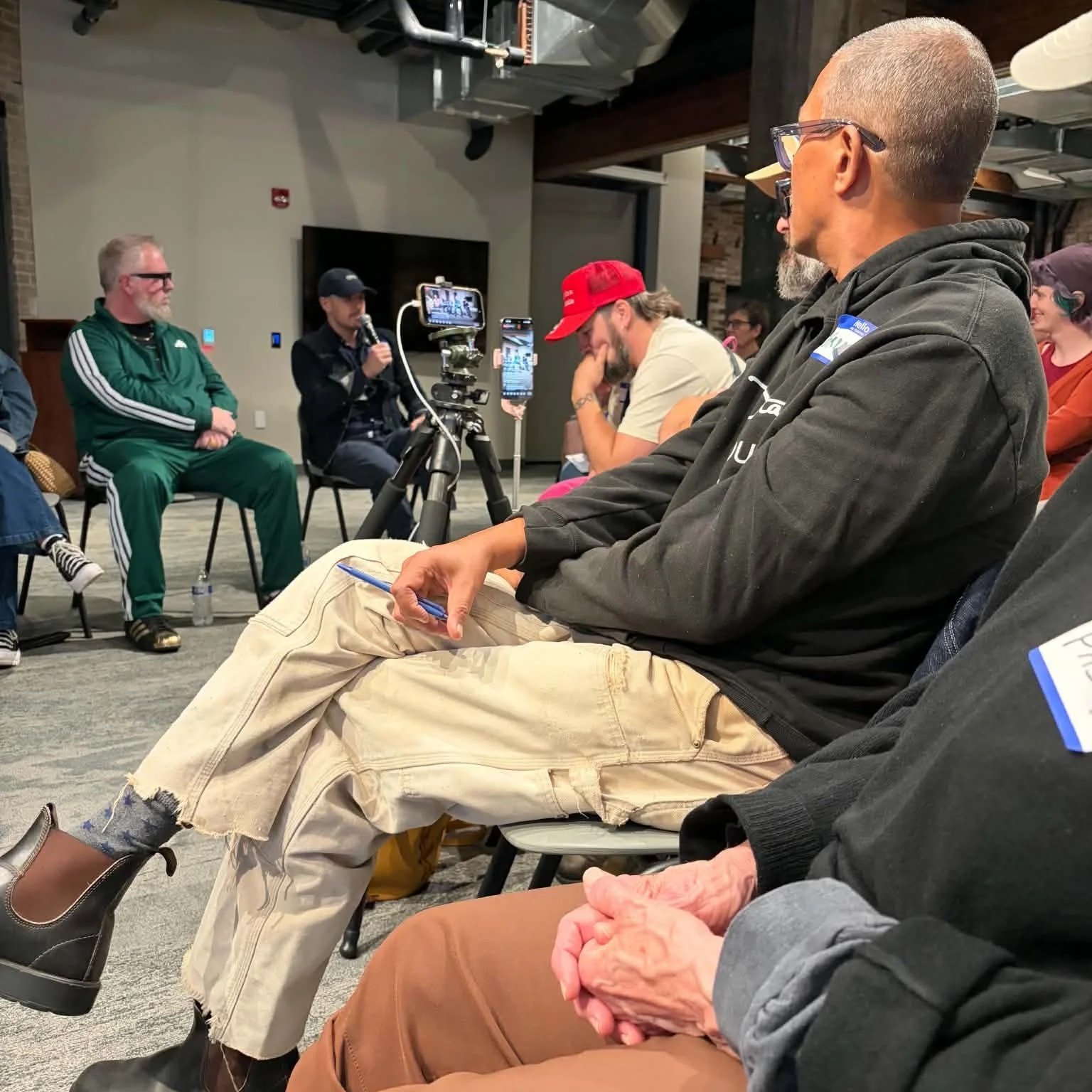 A group of people attending a panel discussion or briefing, with one person speaking into a microphone, and others sitting and listening, some taking notes or looking at their phones.