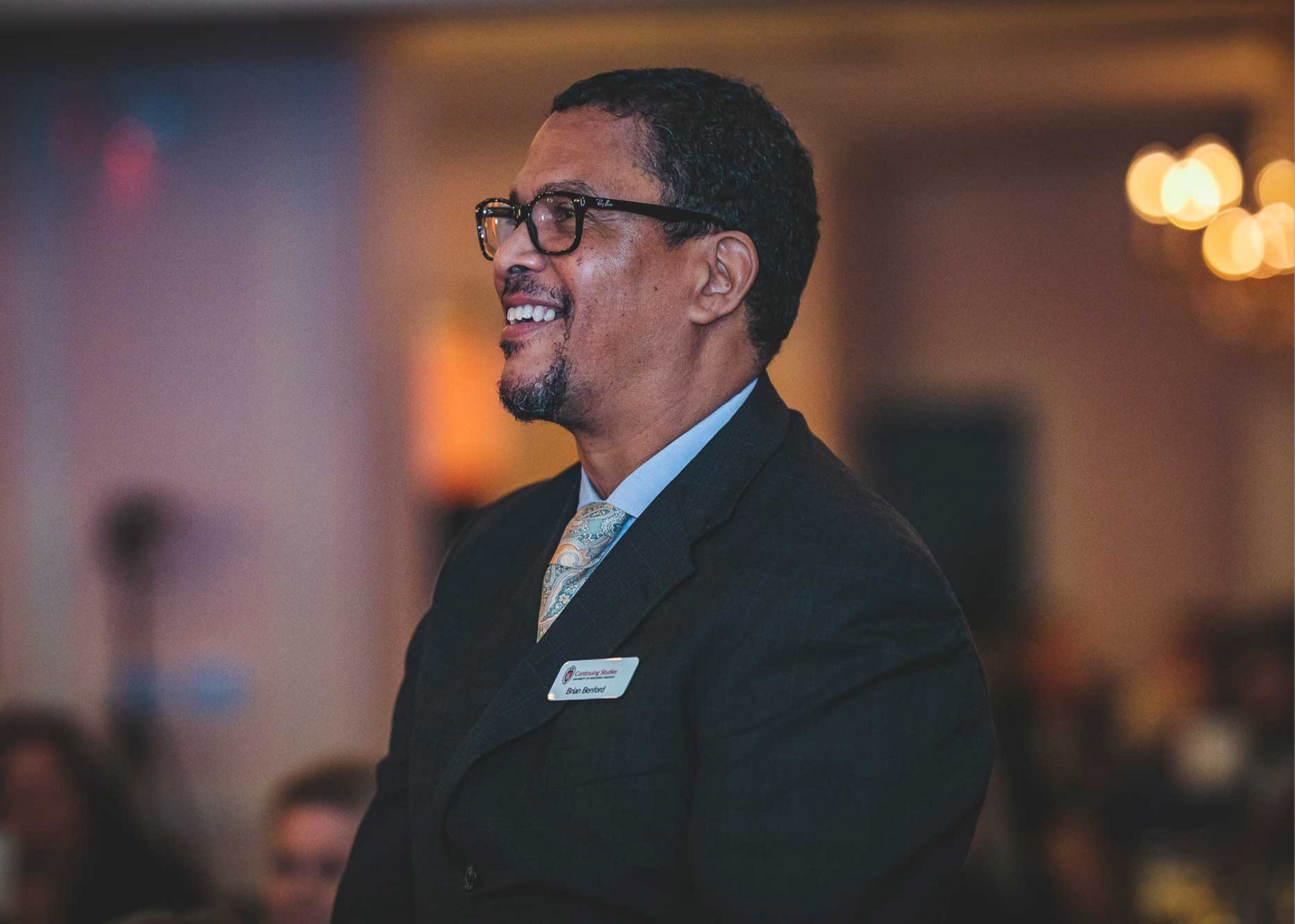 A man with dark curly hair and glasses, dressed in a dark suit with a dress shirt and a patterned tie, smiling at an indoor event.
