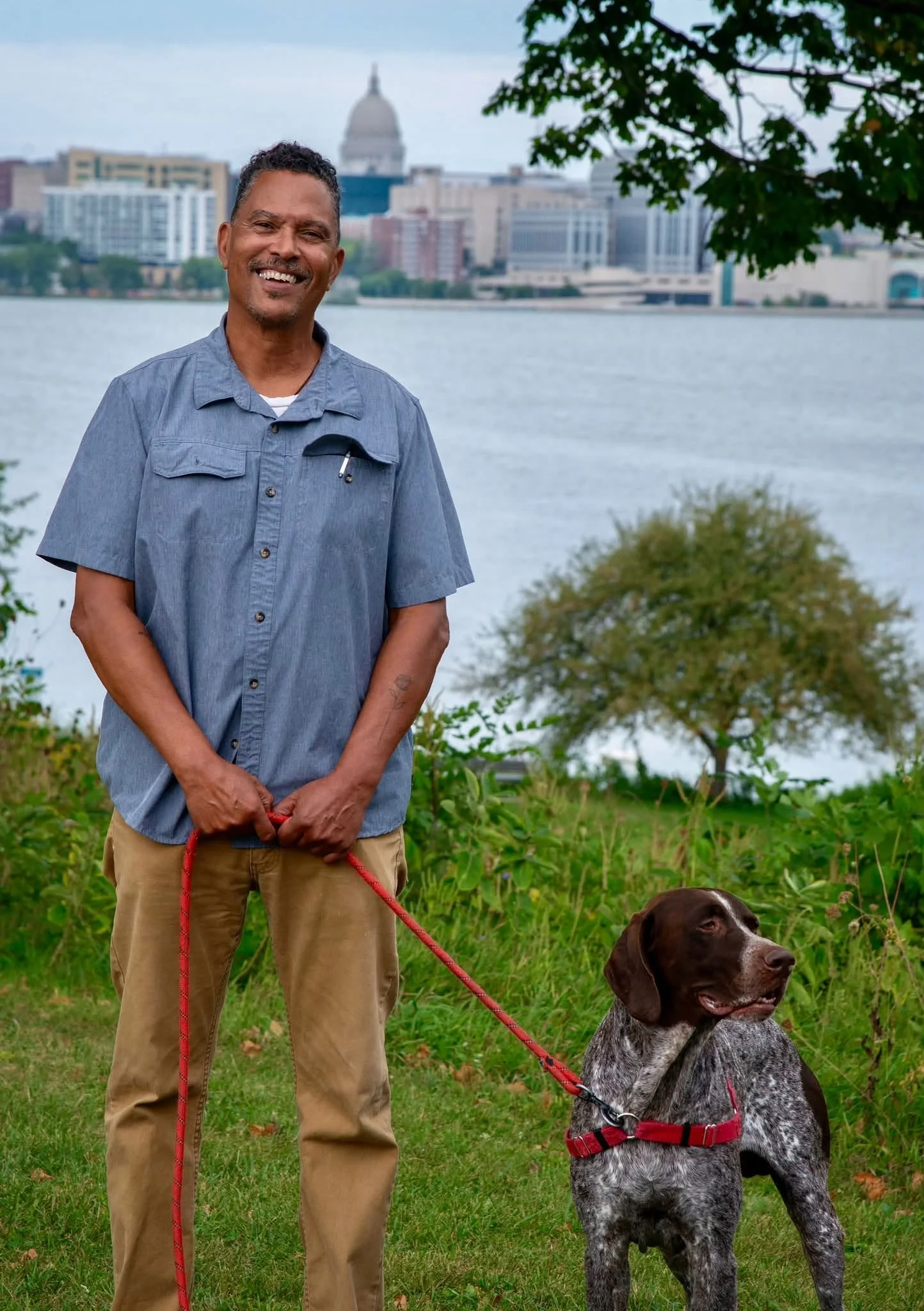 A man with short curly hair, smiling, standing outdoors by a body of water with a city skyline in the background, holding a leash attached to a brown and white dog with a red harness, sitting on the grass under a tree.