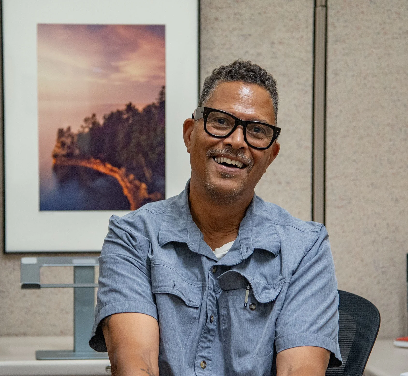 Smiling man with curly hair and glasses sitting at a table in an office, with a framed landscape photograph of a sunset over trees on the wall behind him.