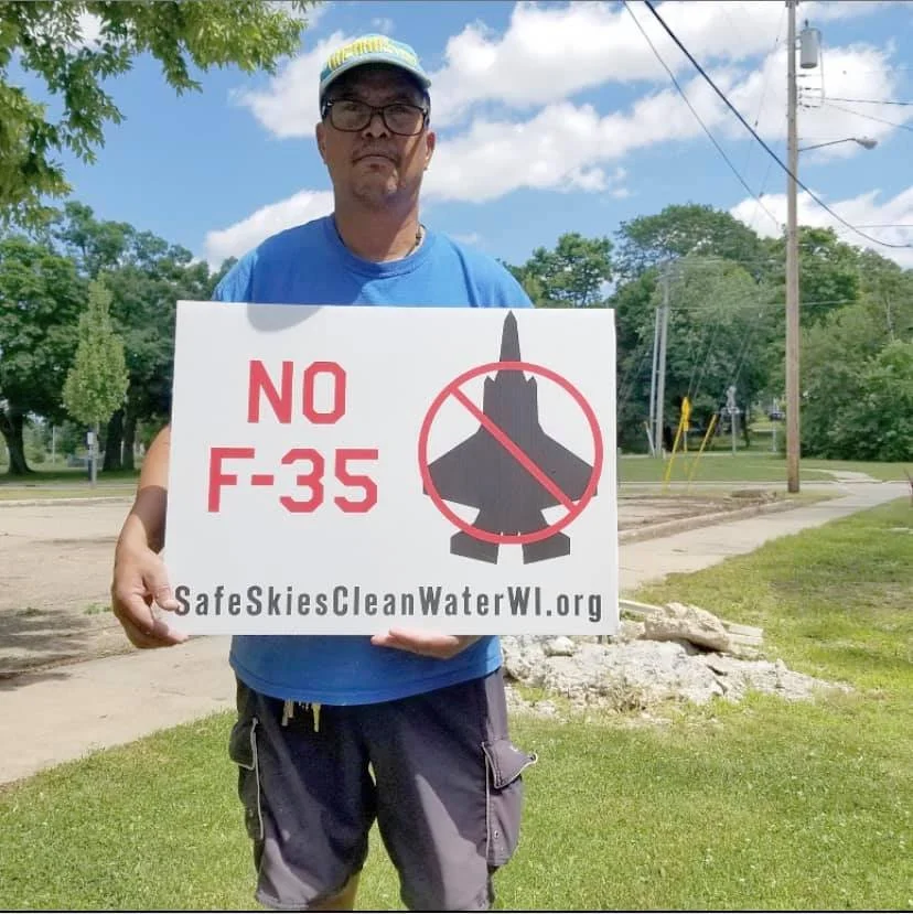 Man holding a sign that says 'NO F-35' with a crossed-out silhouette of a fighter jet and a website URL SafeSkiesCleanWaterWI.org.