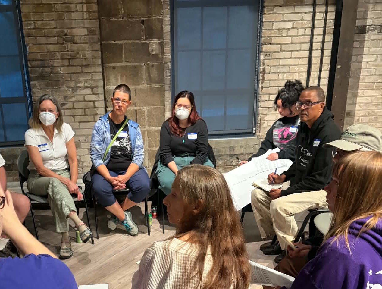 Group of people sitting in a circle during a discussion or meeting, indoors with exposed brick wall and large windows in the background.