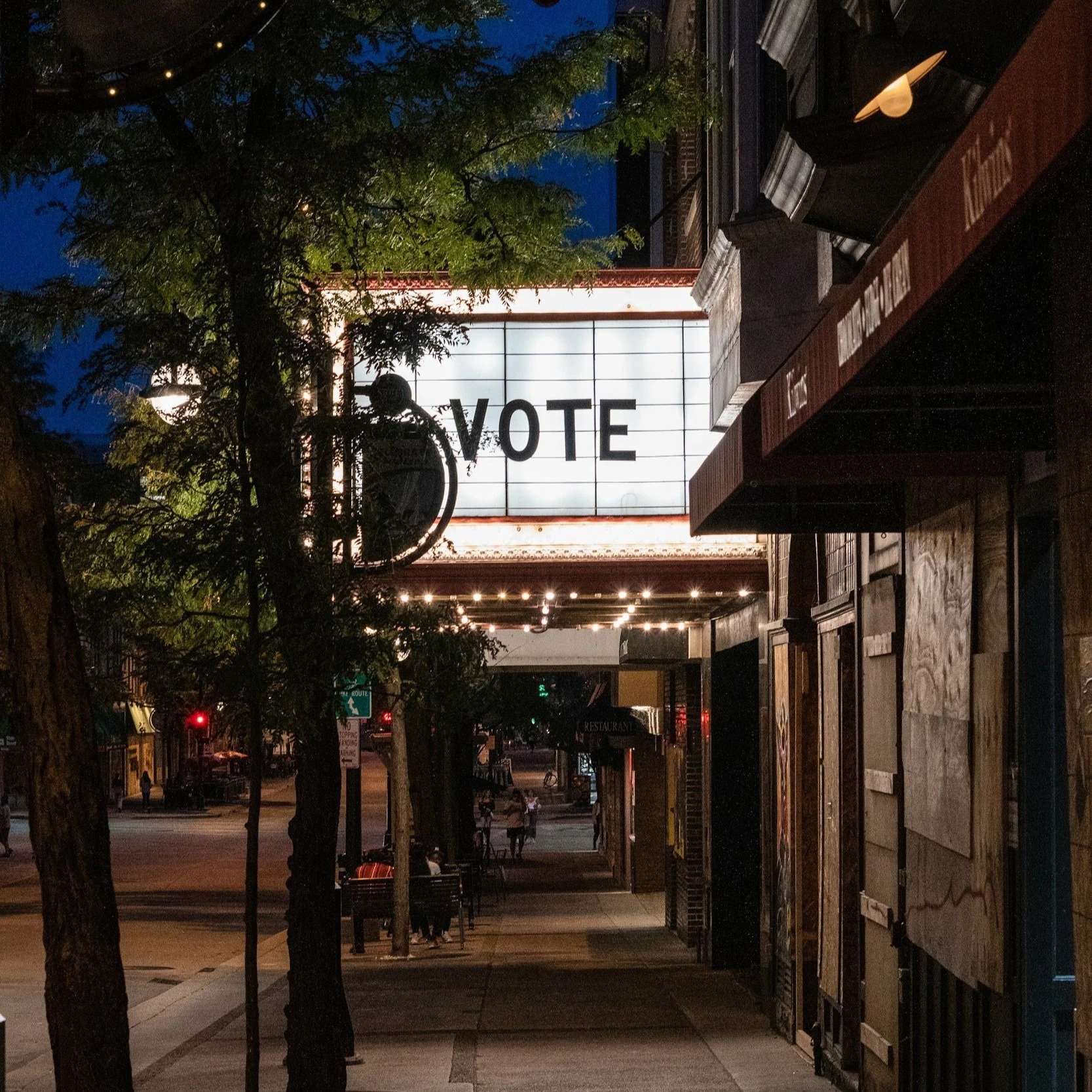 Nighttime street scene showing a bright, illuminated sign with the word 'VOTE' on a building, with trees and sidewalk in the foreground, buildings, and streetlights in the background.