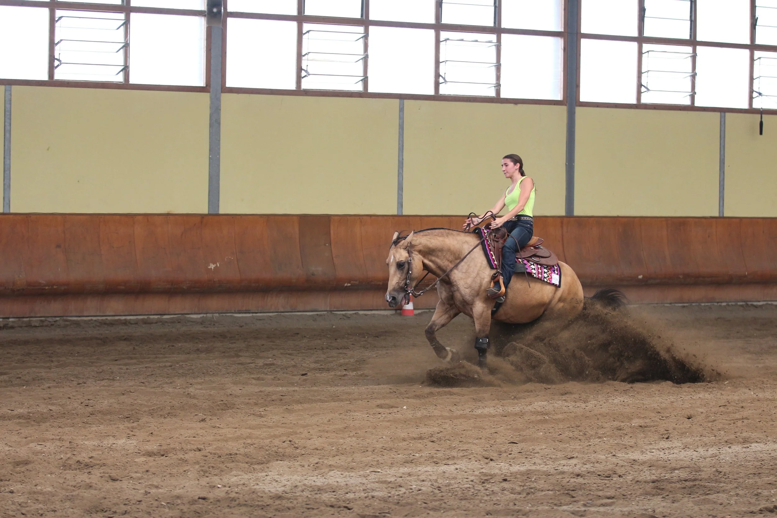 A woman riding a light brown horse in an indoor riding arena, wearing a sleeveless yellow top and jeans.