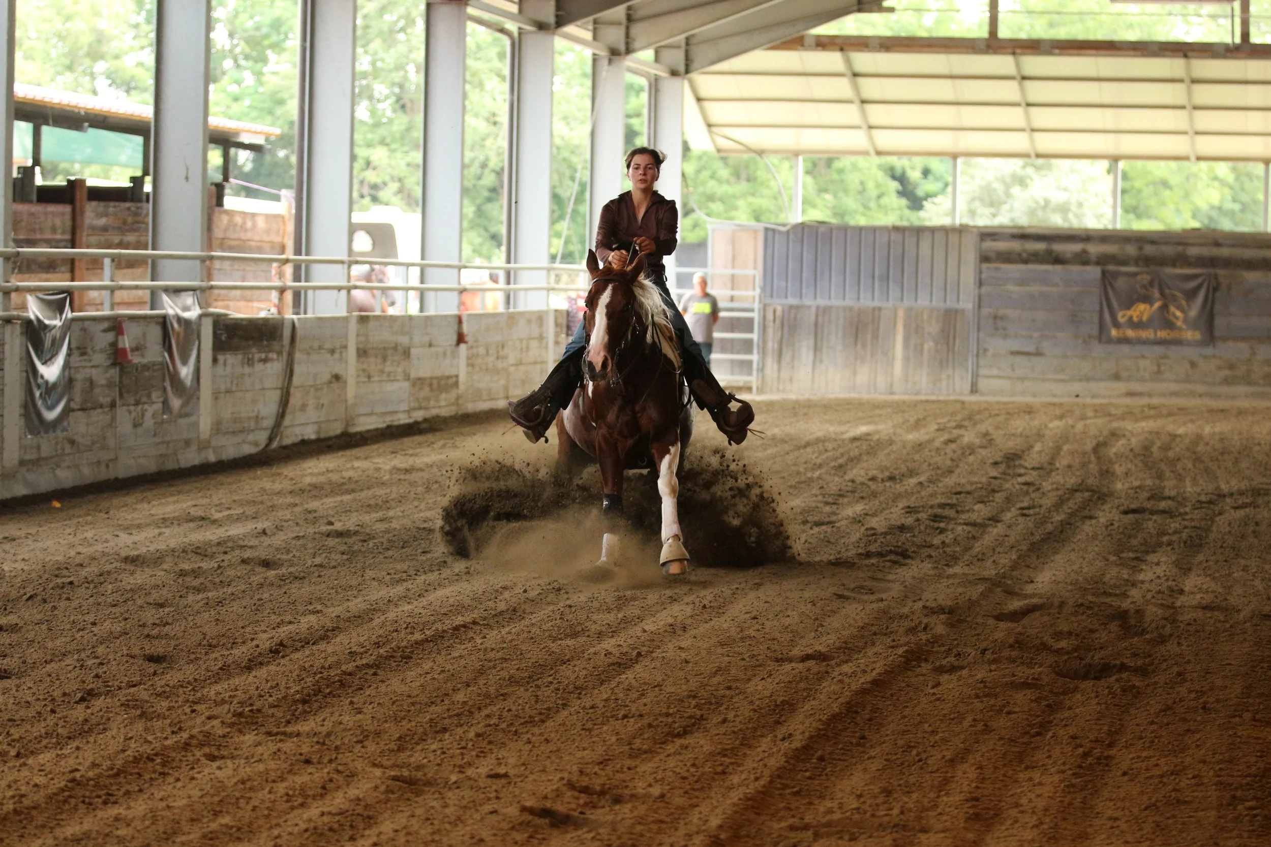 A person riding a horse in an indoor arena with a dirt floor, surrounded by a low wooden fence, and green trees visible through the windows.