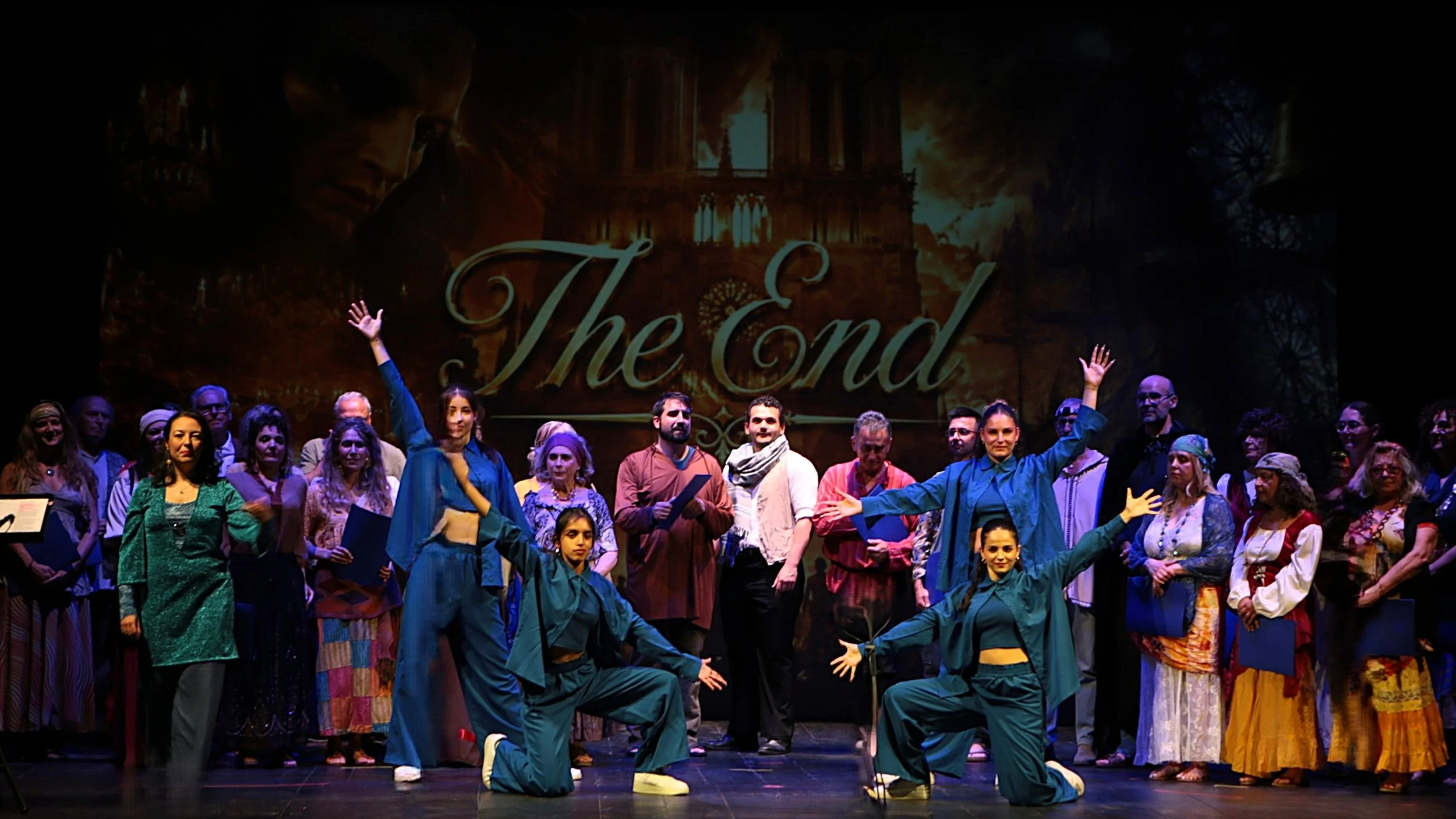A group of performers on stage, some kneeling and some standing, in front of a projector screen with the words 'The End' and an image of a Gothic cathedral. The performers are dressed in colorful costumes, and some are waving or posing with arms outstretched.