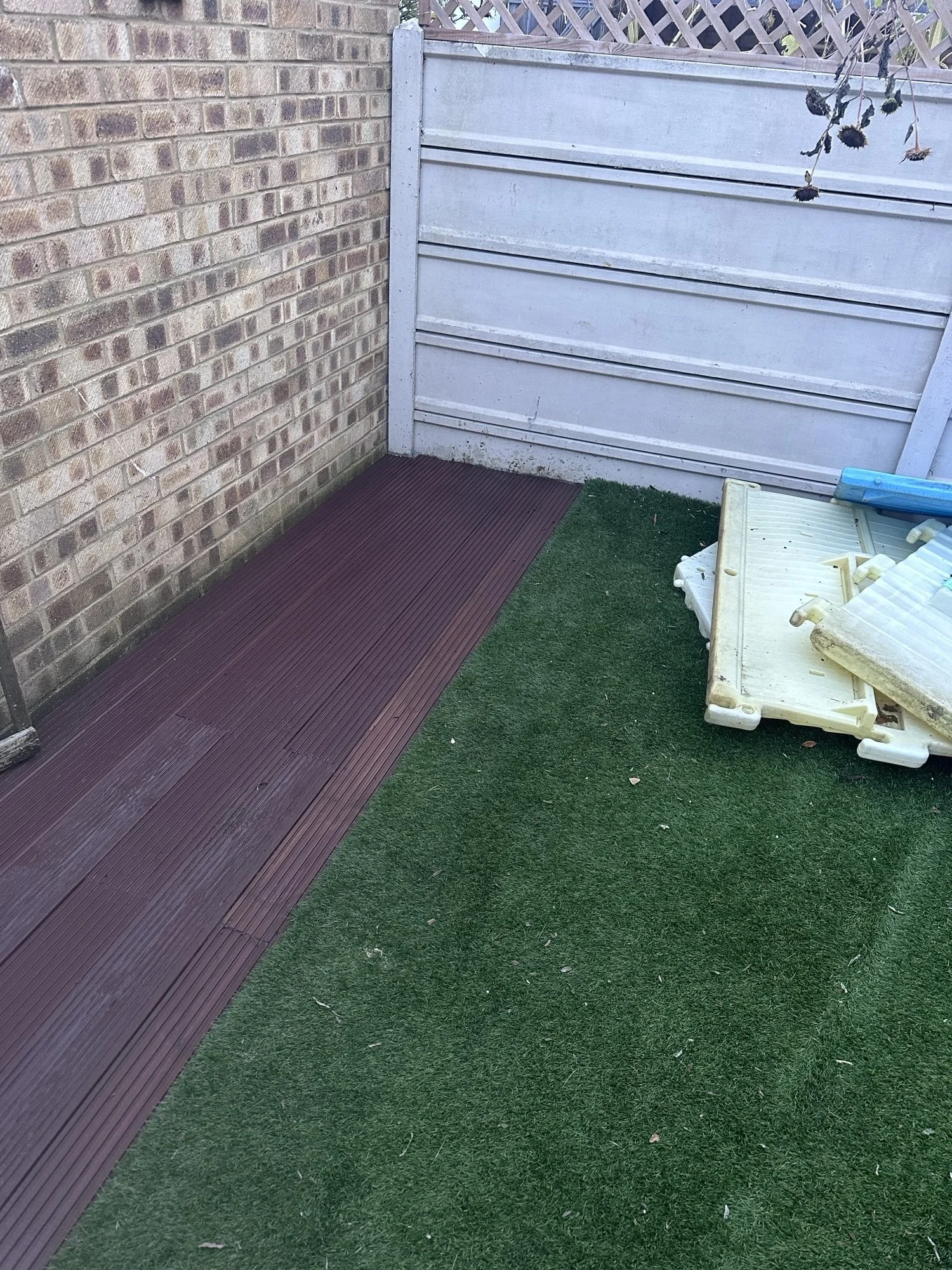 A small outdoor area with a red rubber mat, artificial grass, and a white fence. There are discarded white plastic pieces and a blue object on the ground.